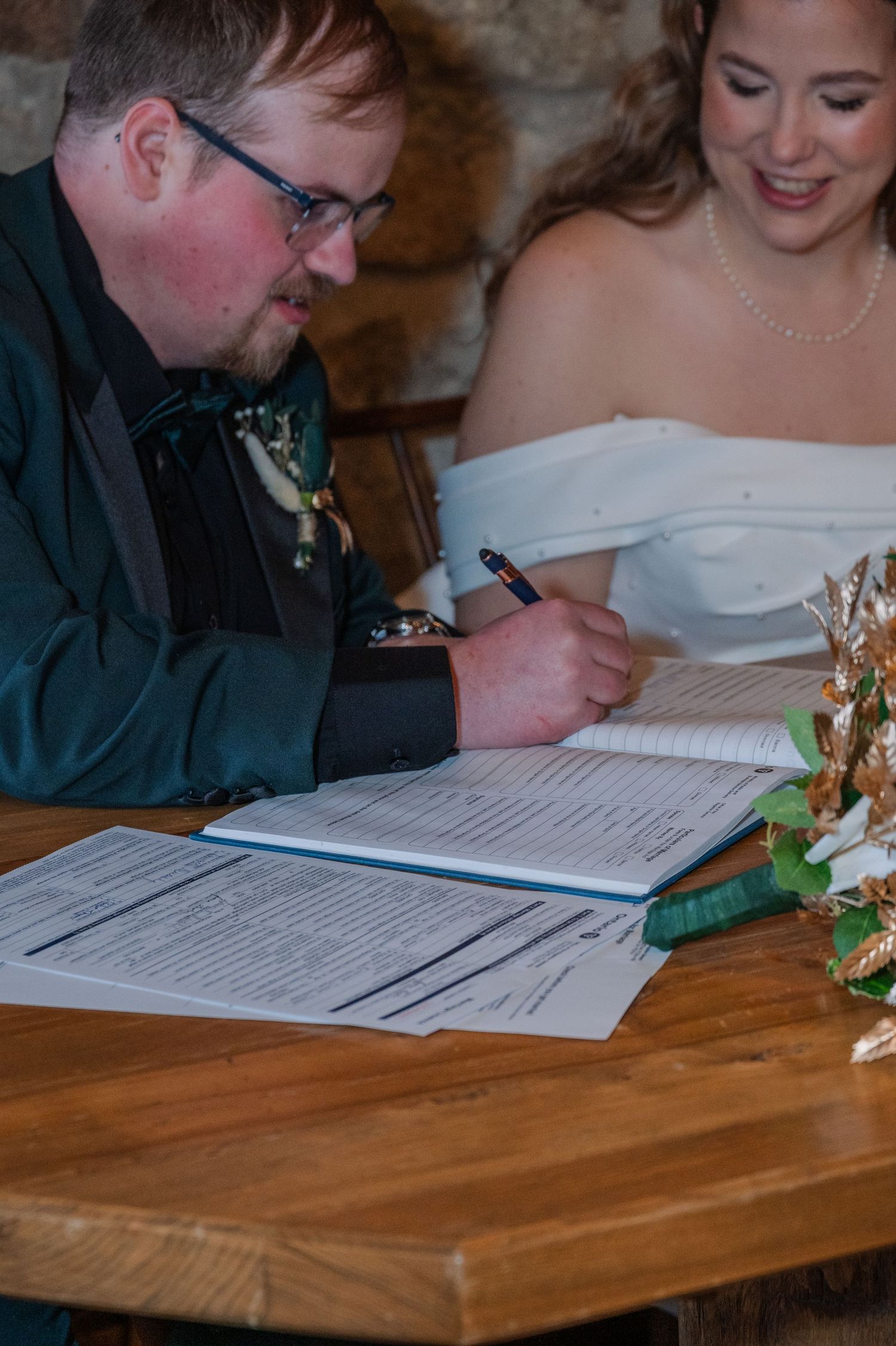 Groom signing wedding marriage certificate at wooden table with bride in white off-shoulder dress smiling behind him.