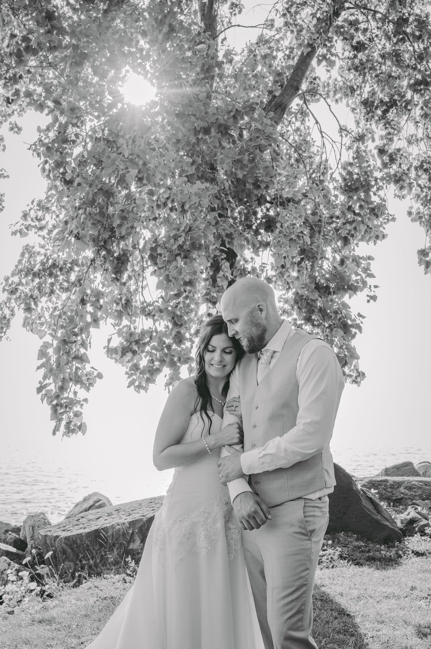 Romantic black and white portrait of couple embracing under sunlit tree by the water.