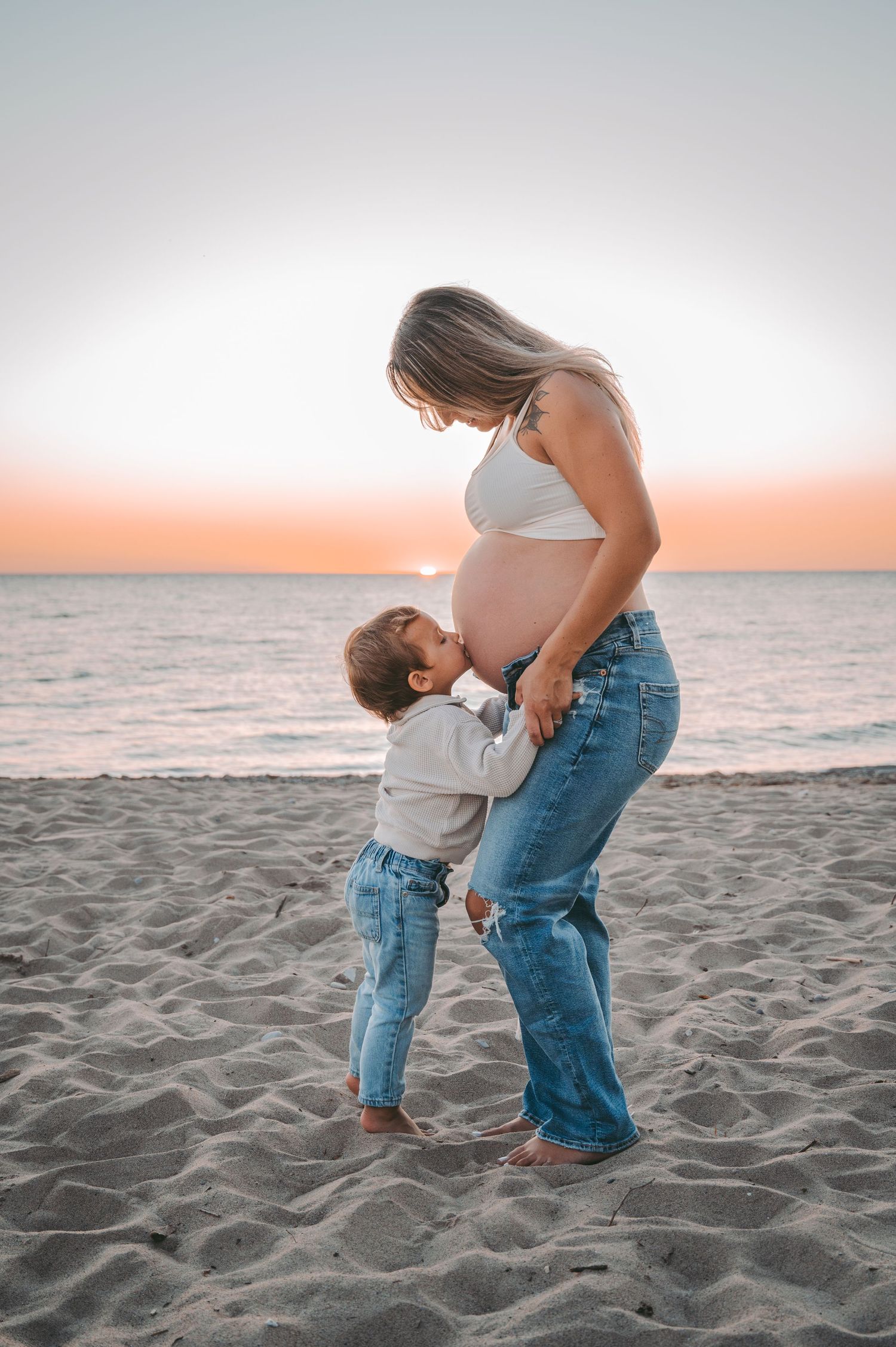 Mother and child share tender moment on beach at sunset in casual denim attire.