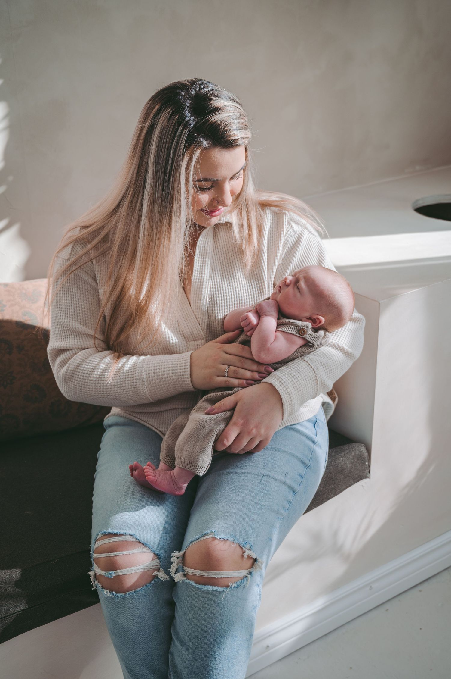 Parent in cream sweater and ripped jeans holding baby while sitting by window in natural light.