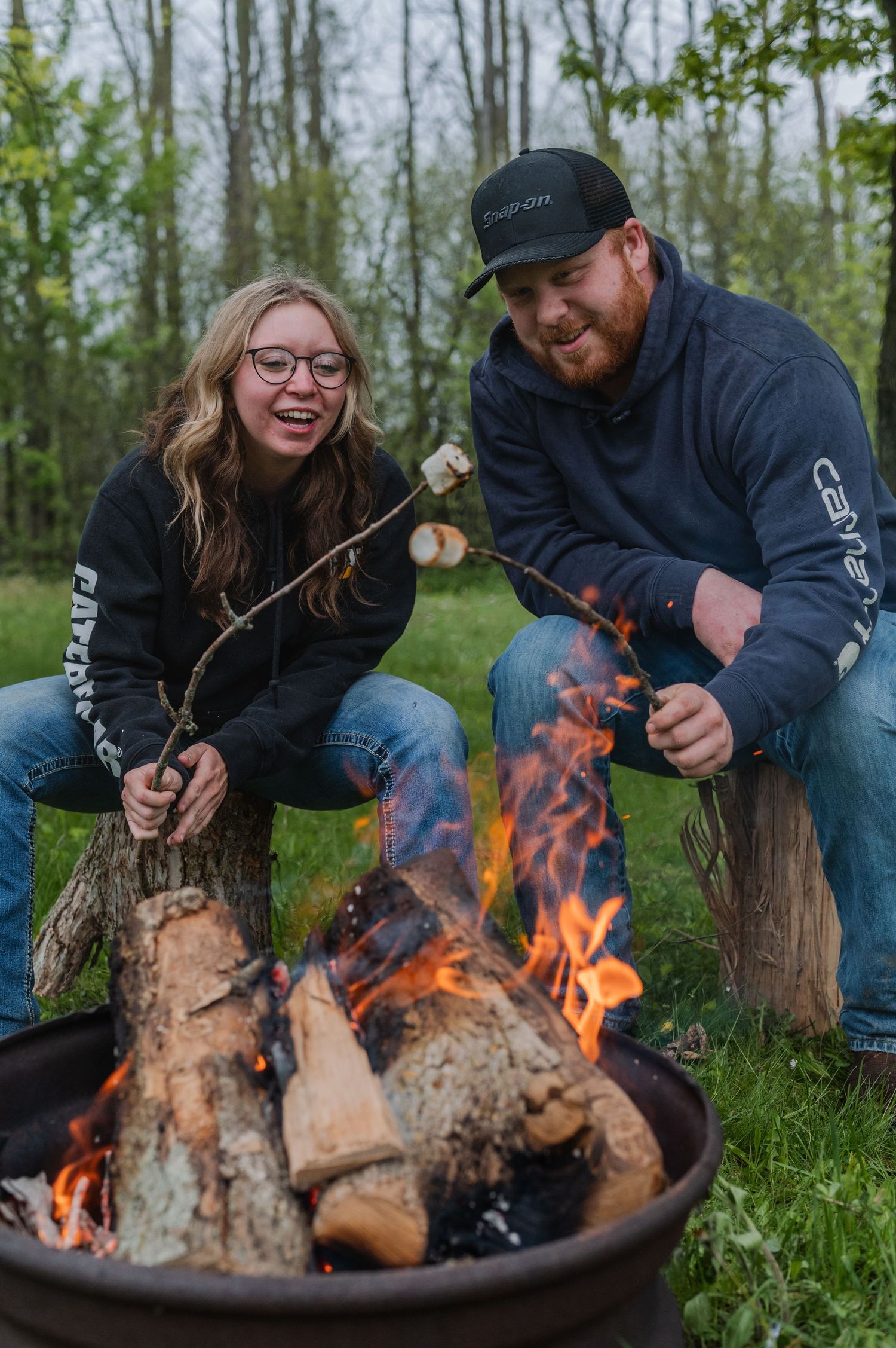 Two people roasting marshmallows over a campfire outdoors, smiling and wearing matching dark hoodies.