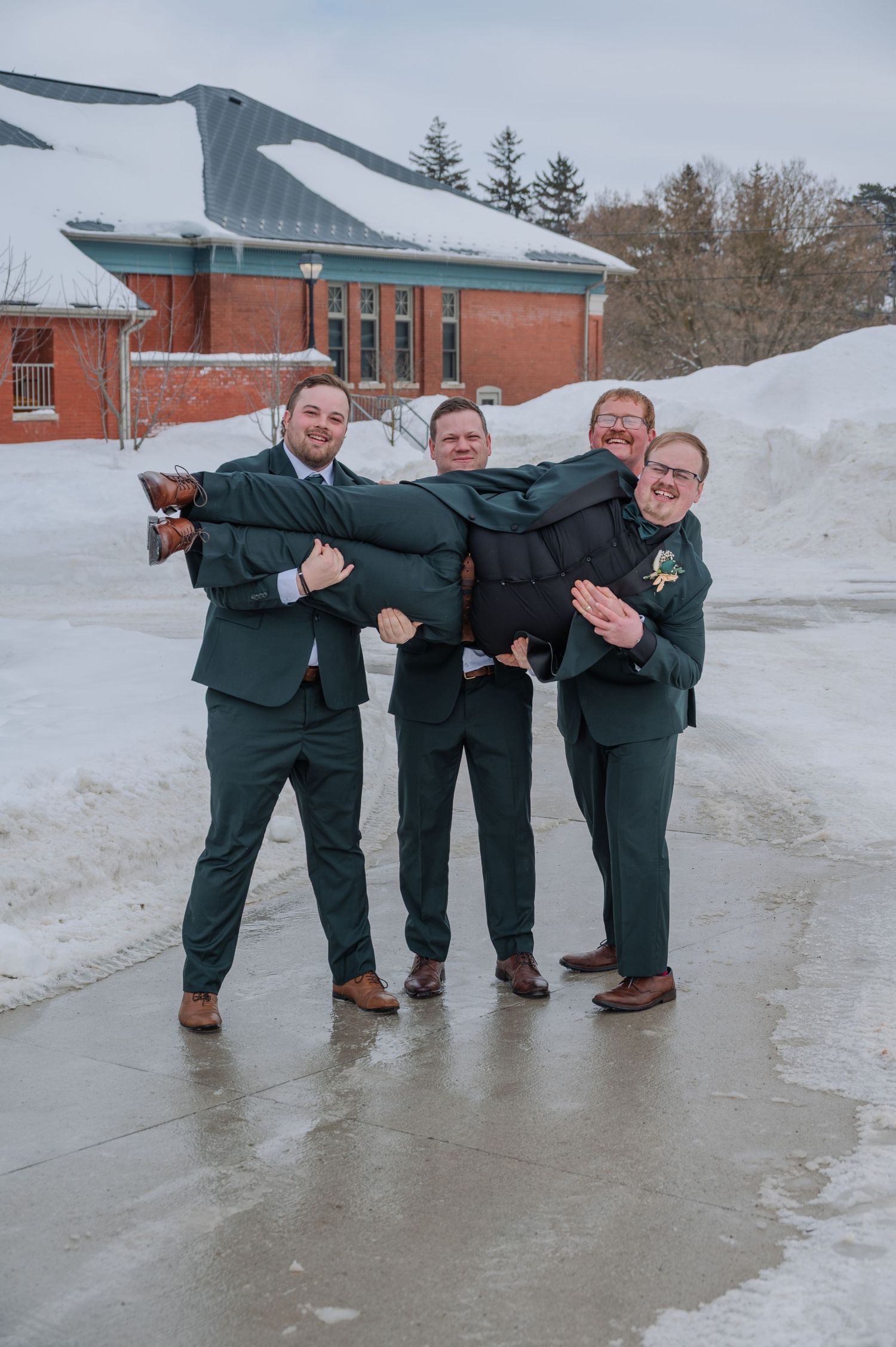 Four men in dark green suits pose playfully outdoors in winter, carrying one man horizontally near a snow-covered brick building.