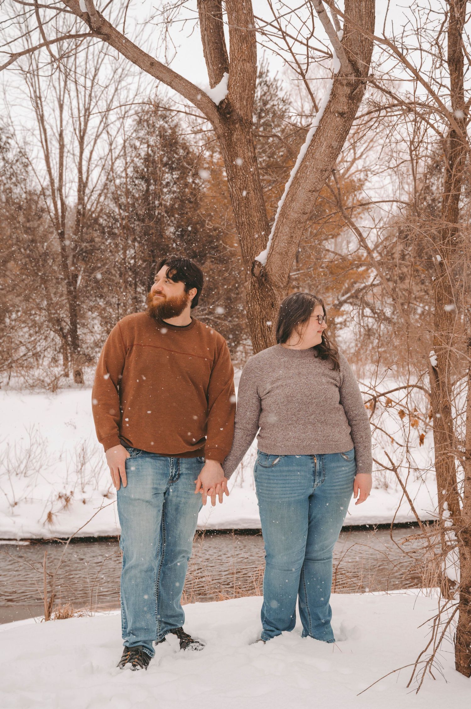 A couple holding hands while standing by a snowy creek with bare winter trees behind them.