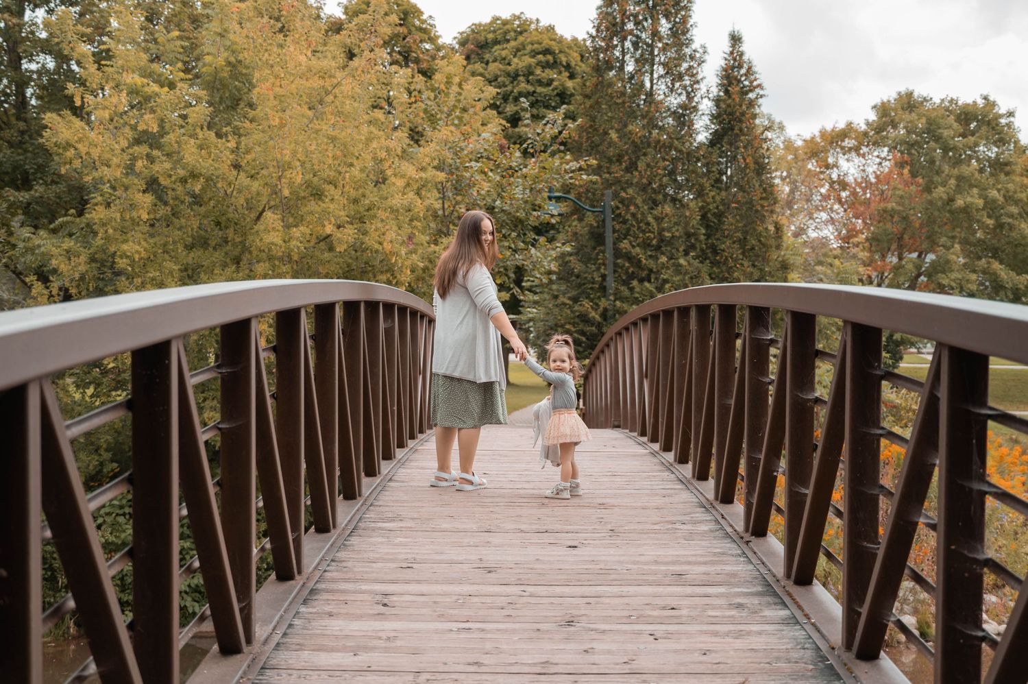 Parent and child walking hand in hand across a wooden bridge surrounded by autumn trees.