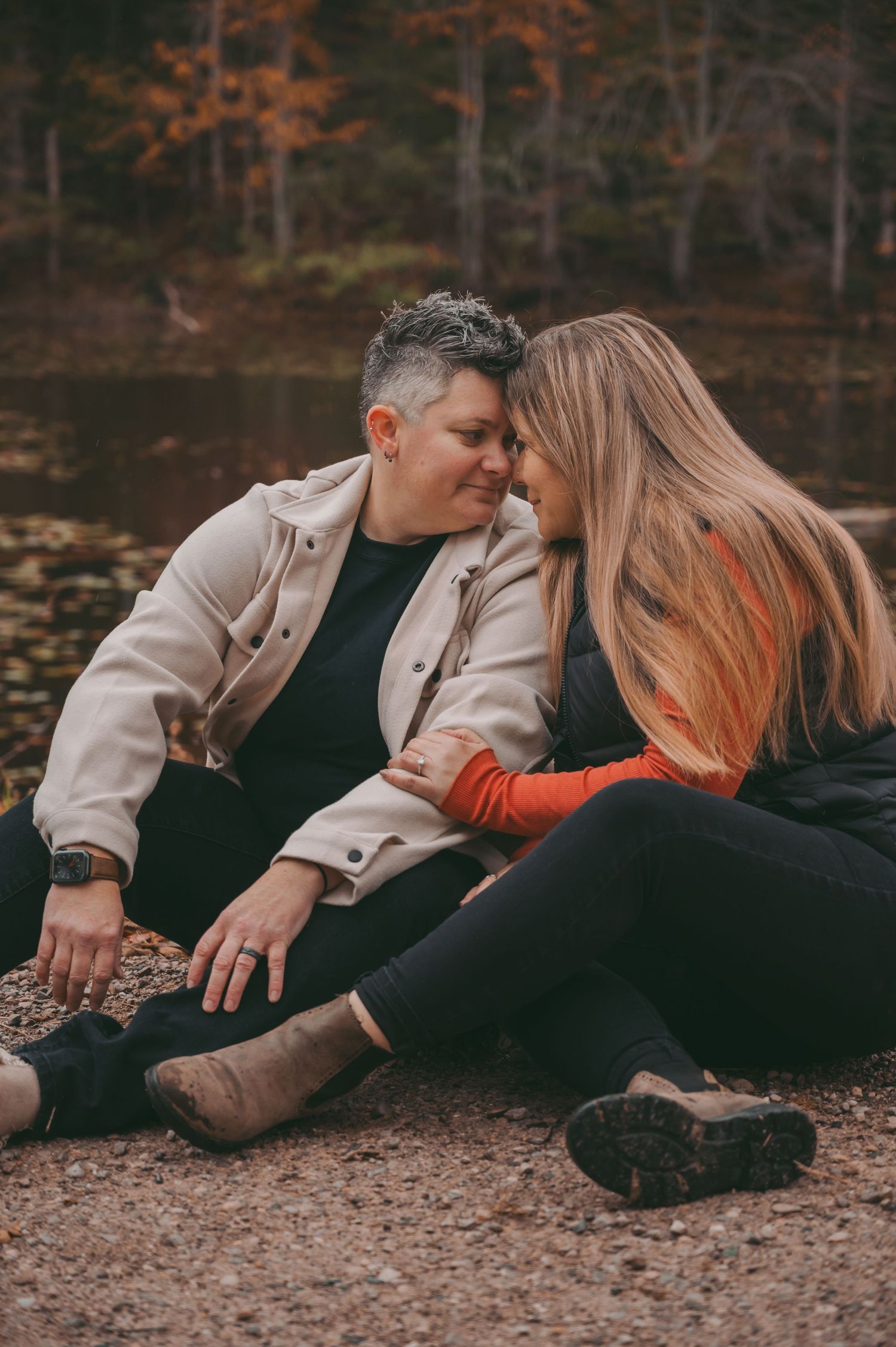 A couple sitting close together by a lake surrounded by fall foliage.