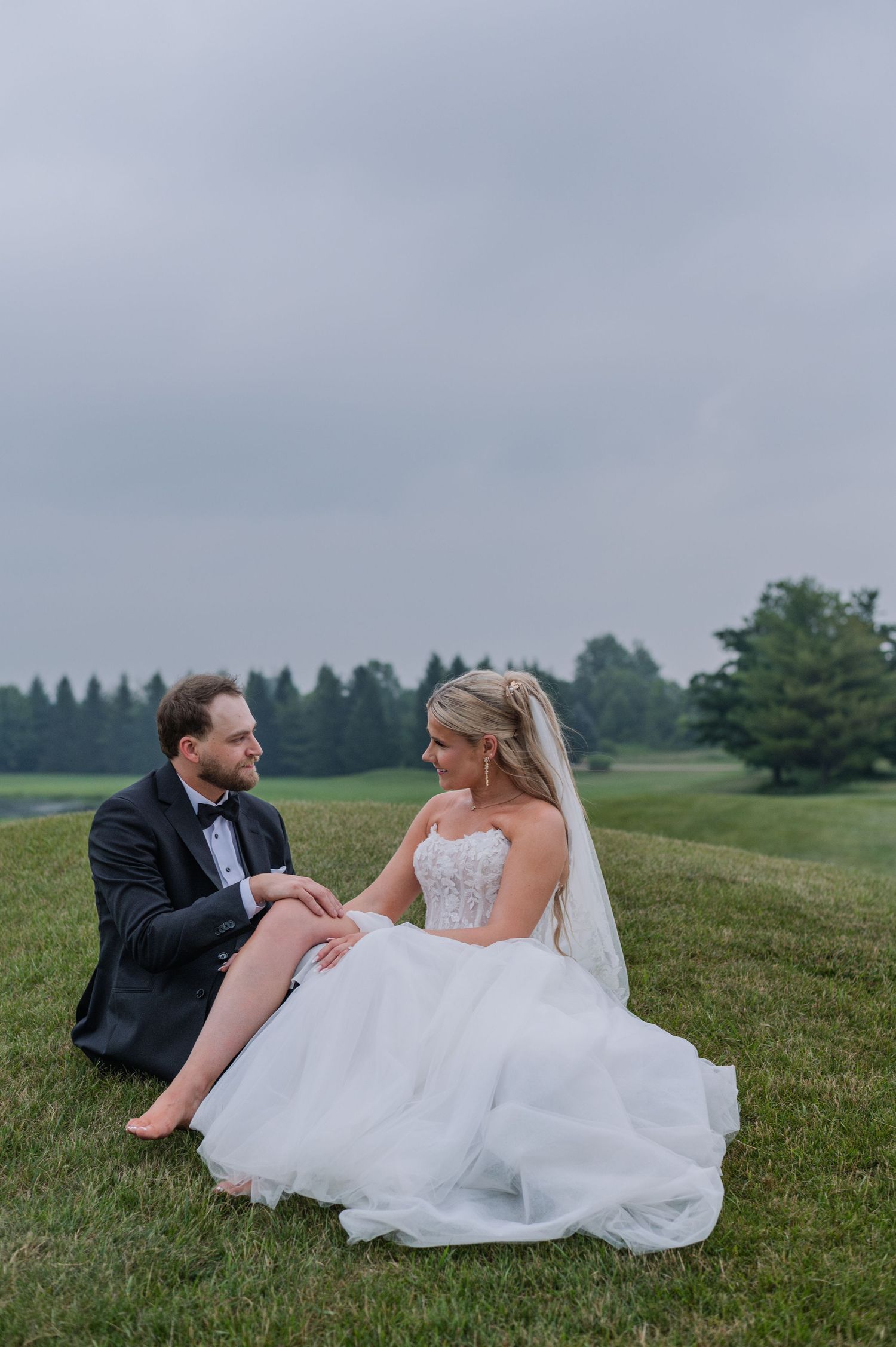 Bride and groom sitting on a lush green hillside in wedding attire, sharing an intimate moment under a cloudy sky.