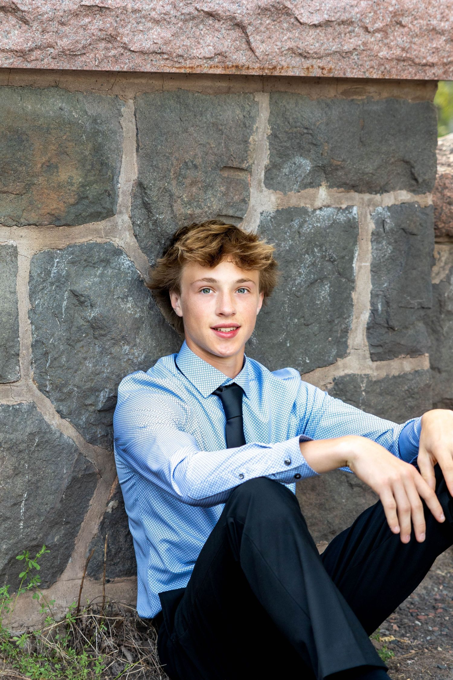 A young person in a light blue dress shirt and dark pants sits casually against a stone wall outdoors.