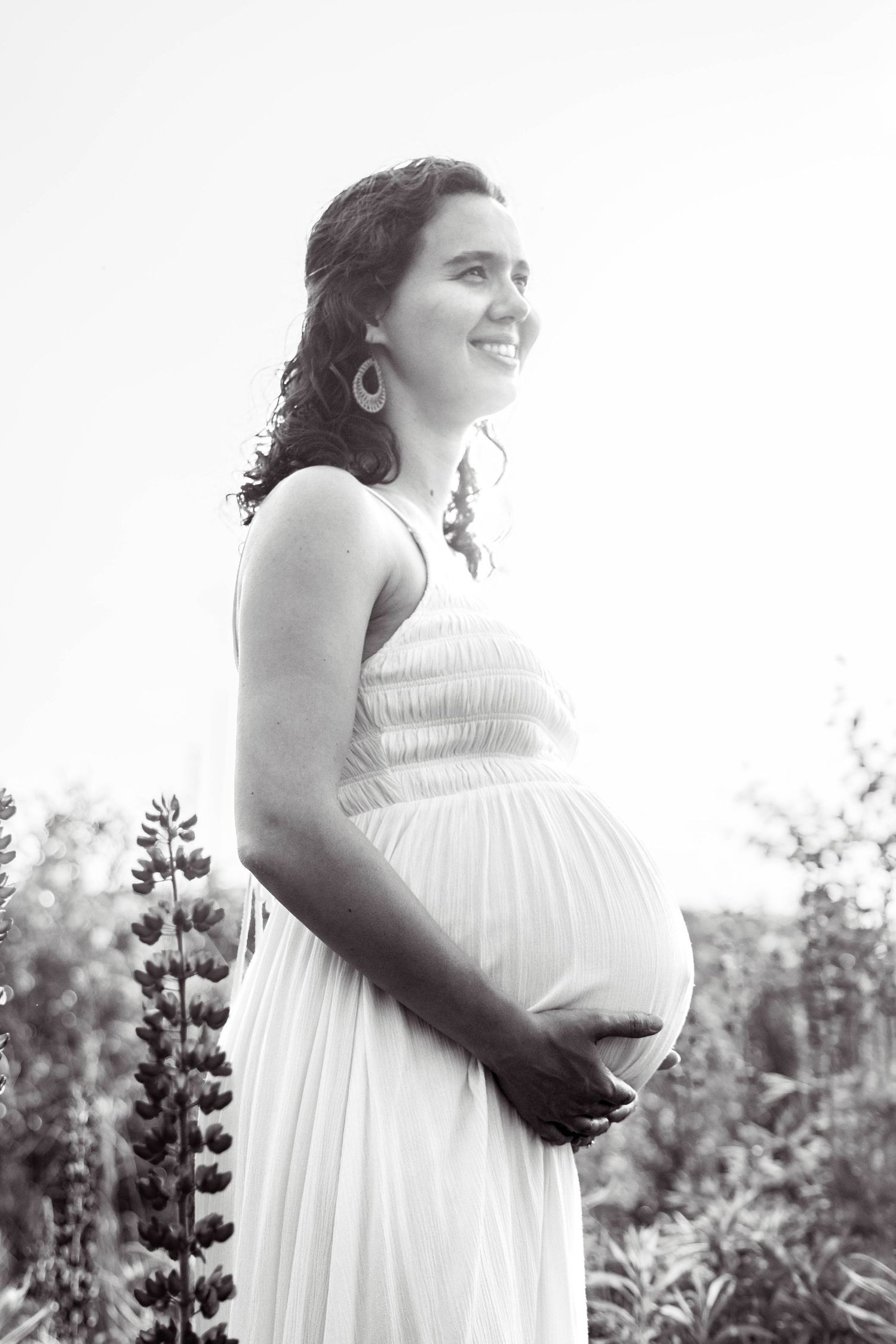 A black and white maternity photo shows a woman in a flowing white dress standing among lupine flowers in a garden.
