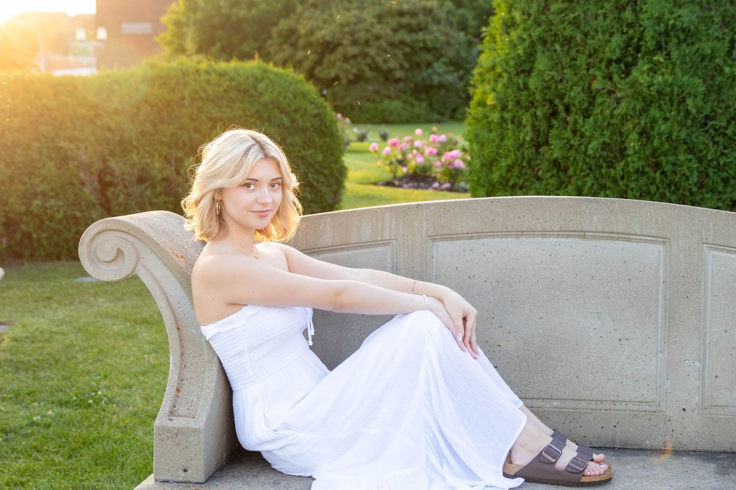 A person in a white dress lounges on a stone bench in a garden during golden hour sunset.