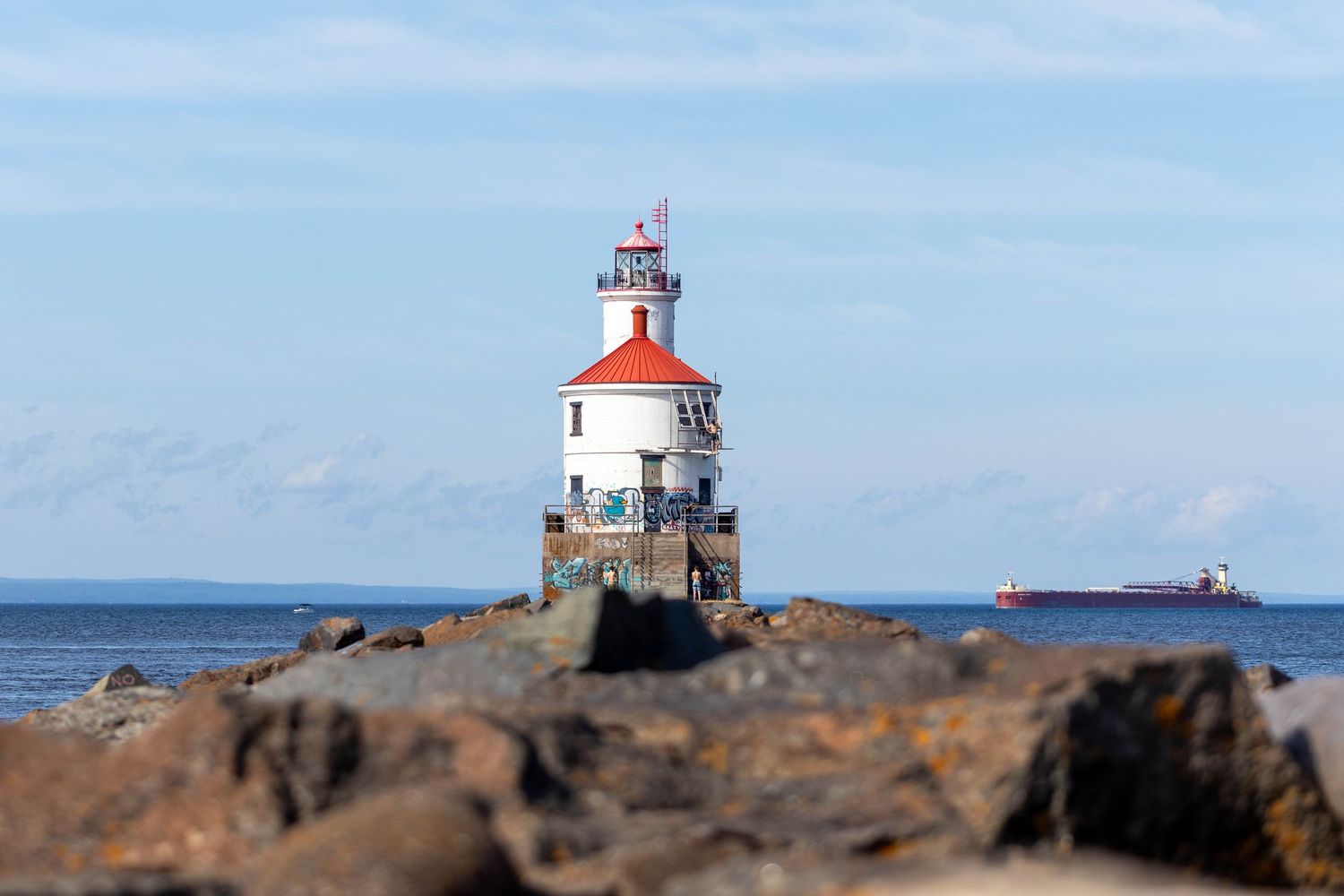 A white and red lighthouse stands on a rocky outcrop in Lake Superior with a cargo ship visible in the distance.