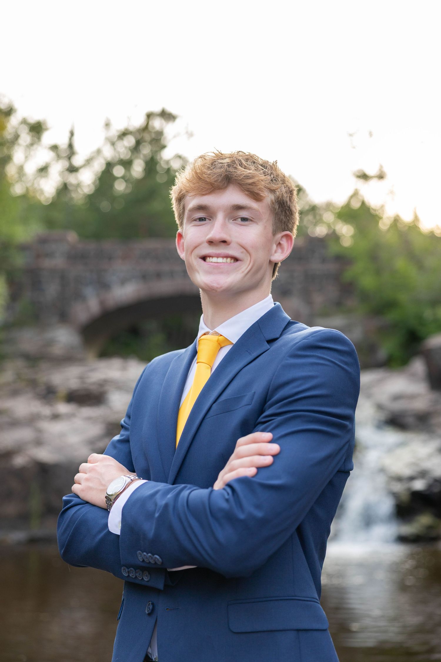 Young man in blue suit and gold tie smiling with arms crossed near a stone bridge and waterfall.