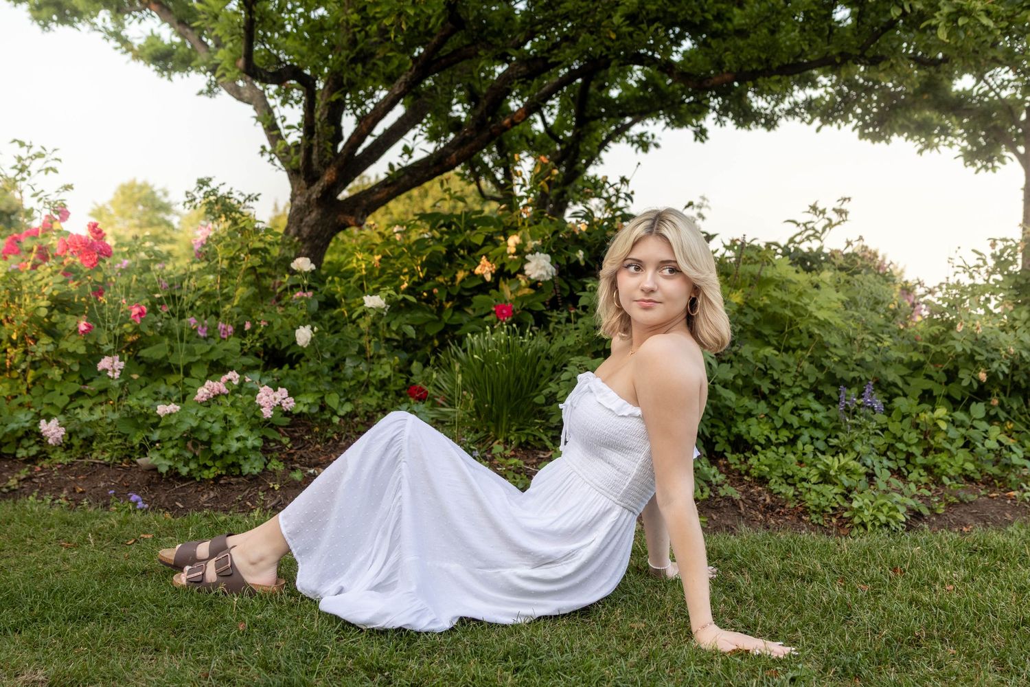 A bride in a flowing white wedding dress poses gracefully on green grass near blooming flowers in a garden setting.