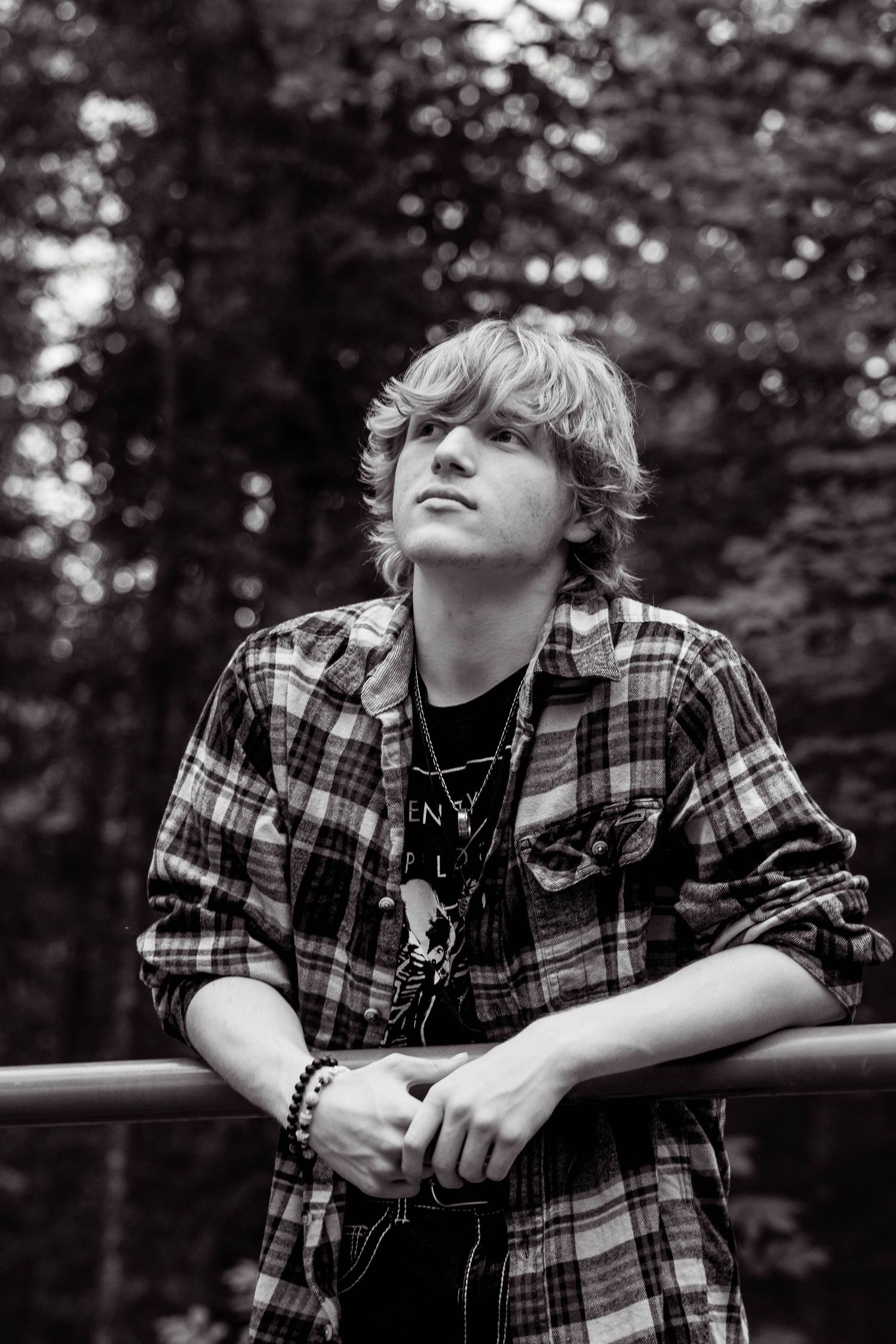 Person in plaid flannel shirt and necklace leans on railing outdoors in black and white photo series.