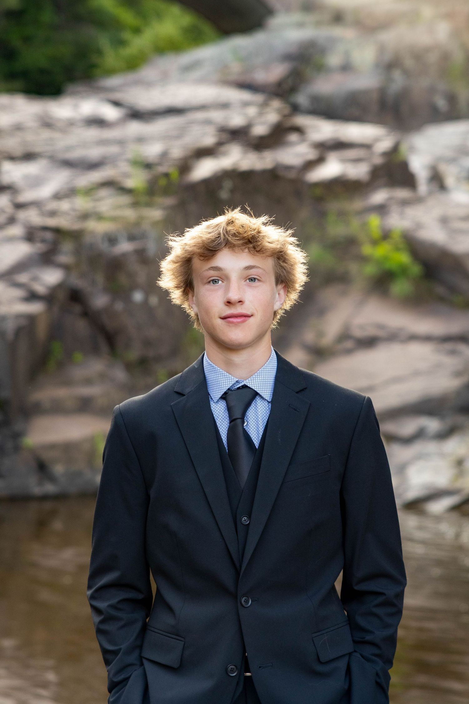 A person in a black suit and tie stands outdoors near a rocky waterfront area.