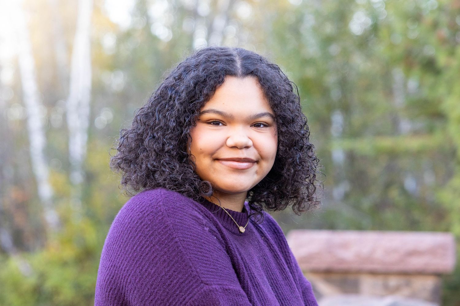 A woman in a purple sweater smiles warmly while posing outdoors with trees in the background.