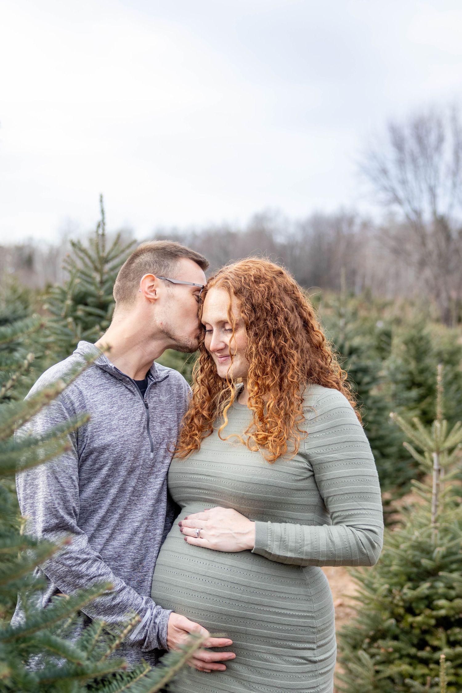 A loving couple shares tender moments between evergreen trees at a Christmas tree farm during a maternity photoshoot.