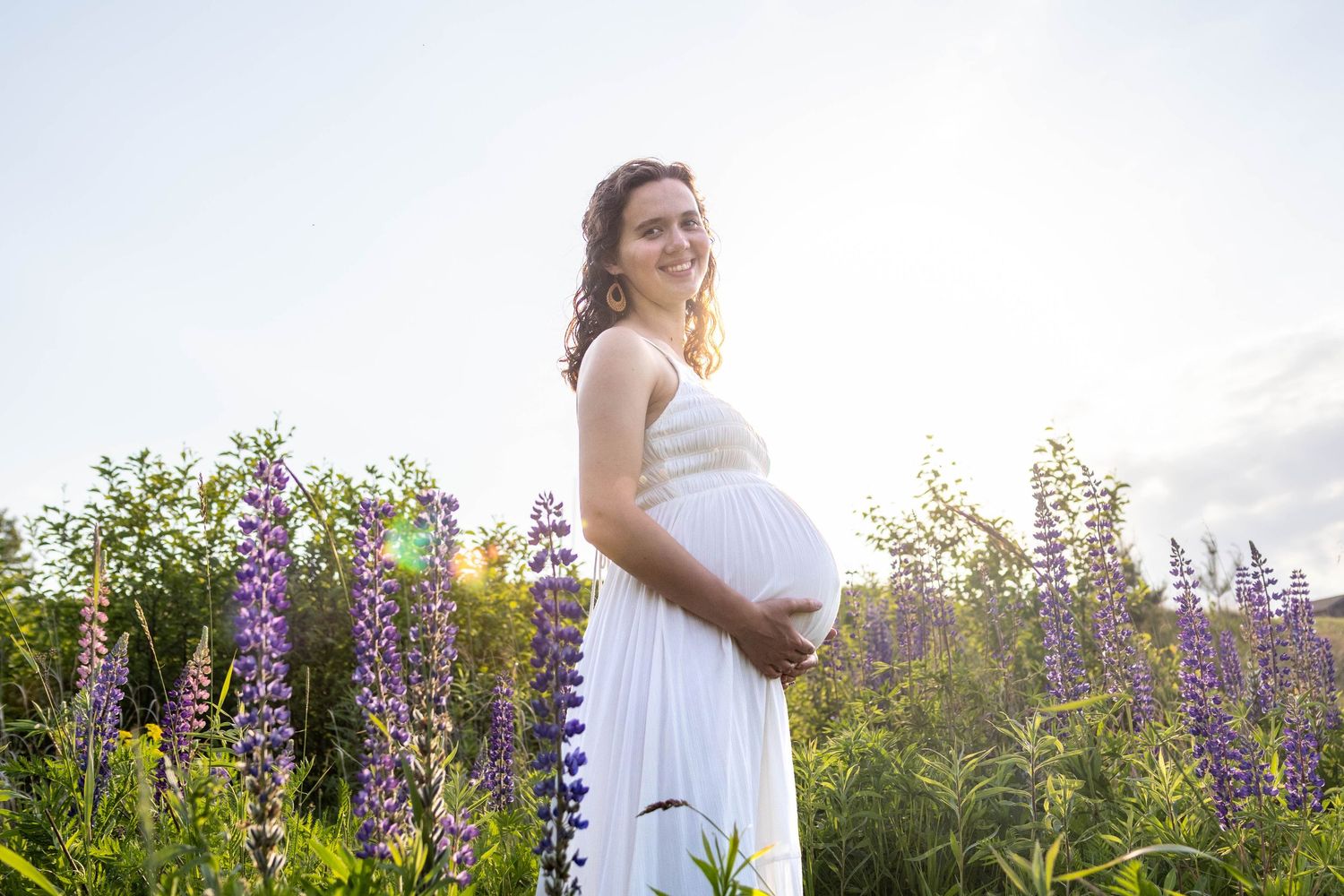 Woman in flowy white dress stands gracefully among vibrant purple lupine wildflowers in a summer meadow at sunset.