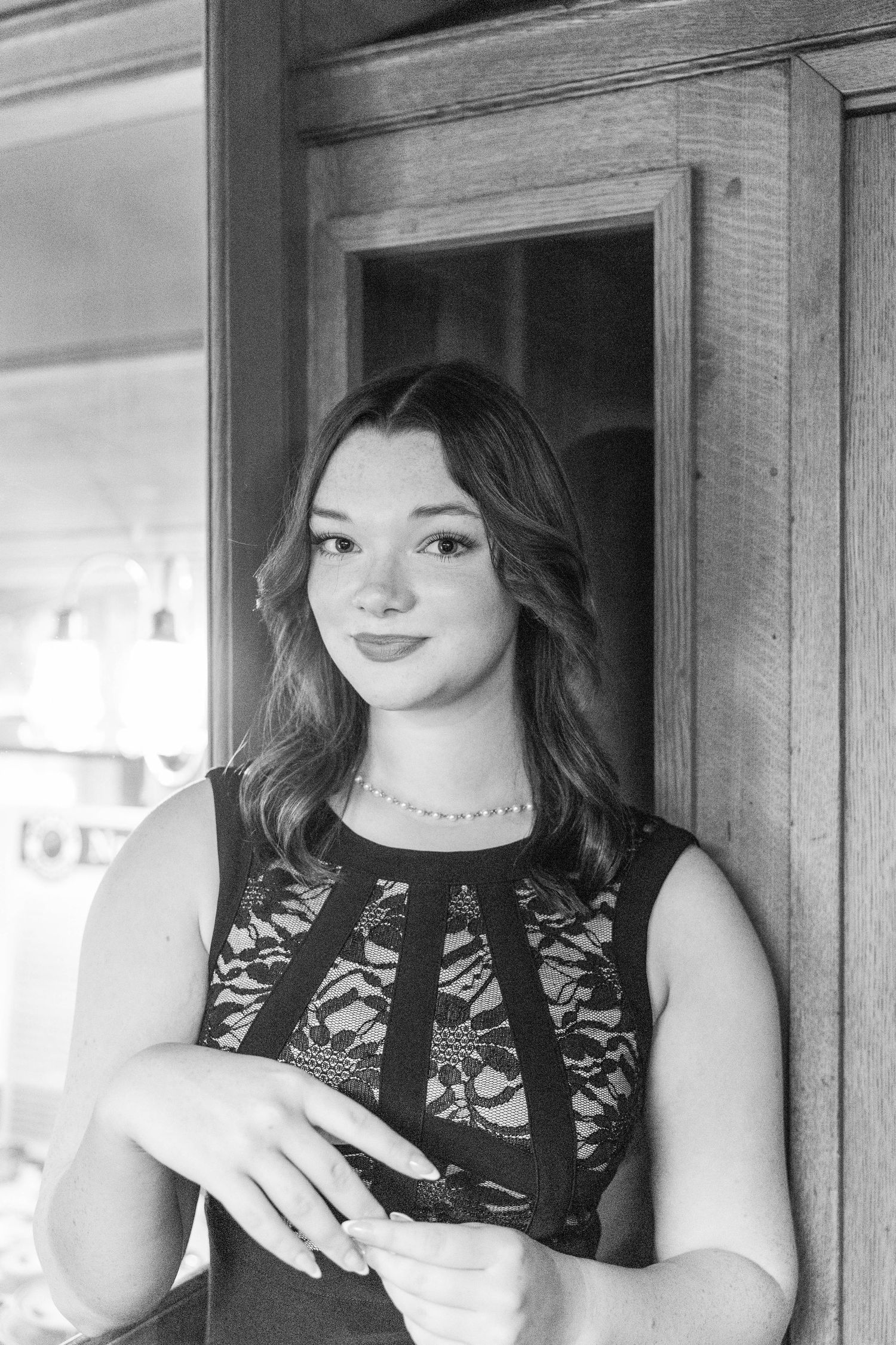 Black and white portrait of a young woman in a lace dress leaning against a wooden door frame.