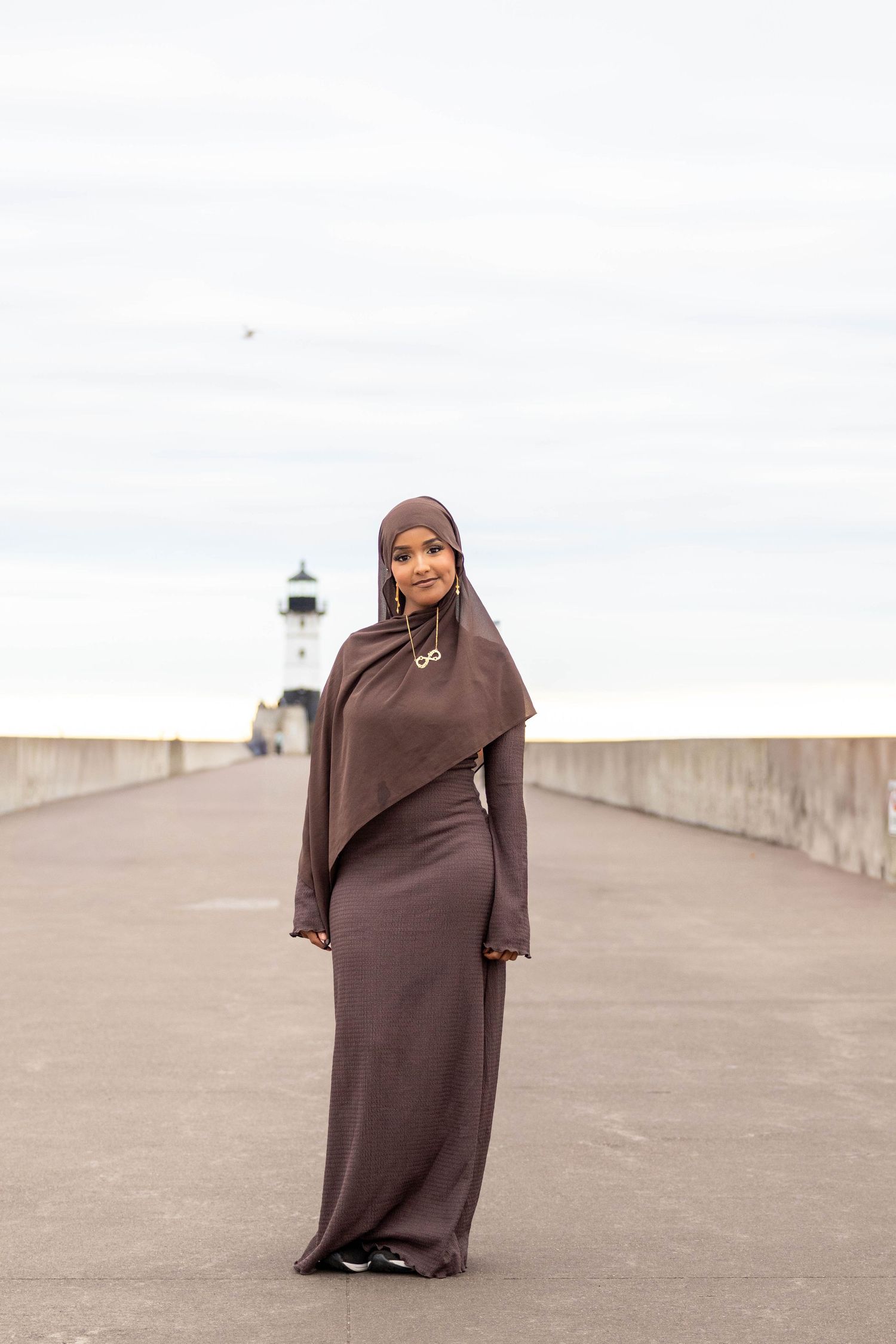 Person wearing a brown abaya stands on a concrete walkway against a cloudy sky.