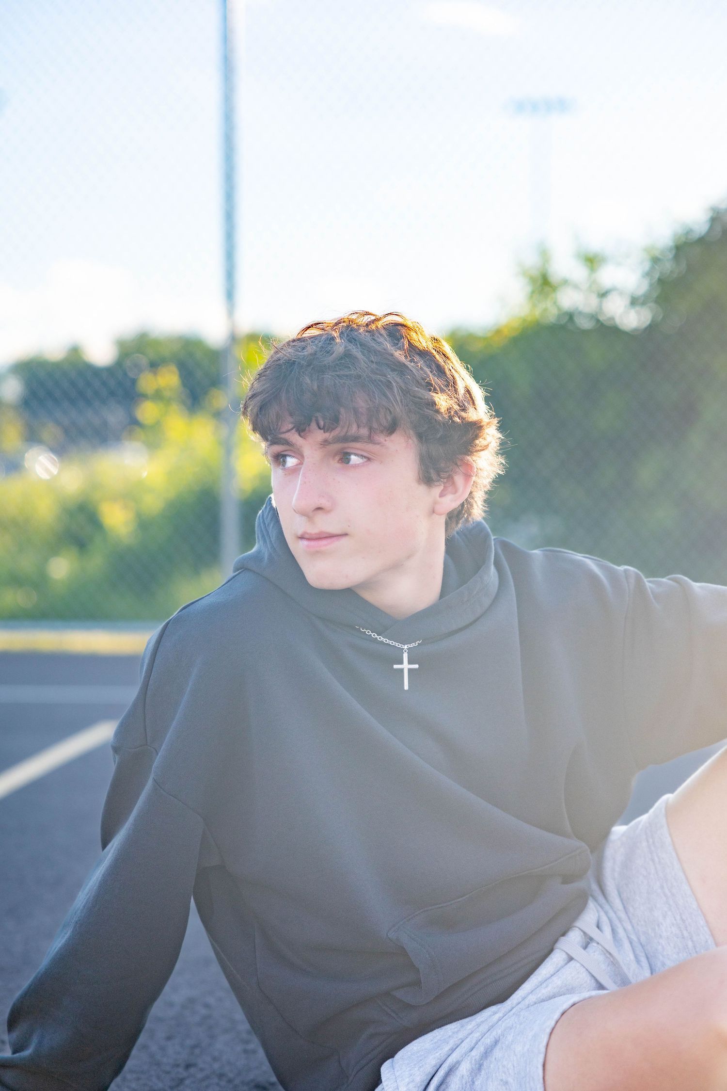 Teen boy in gray hoodie with cross necklace sitting outdoors near chain-link fence on sunny day.
