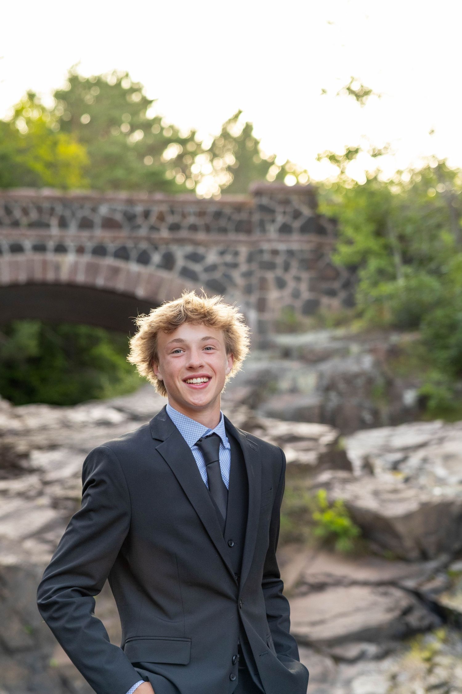 A person in a formal black suit poses outdoors near a stone bridge during golden hour.