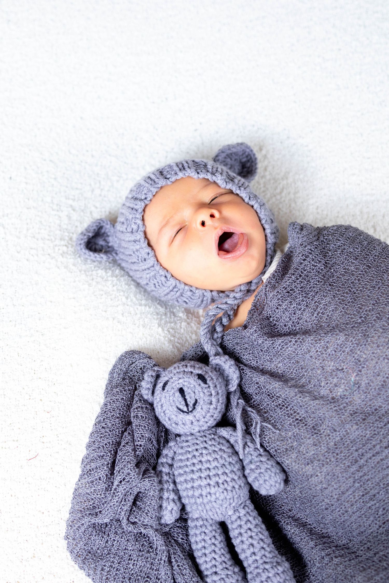 Baby in gray knitted hat and blanket yawning while lying next to matching crocheted teddy bear toy.