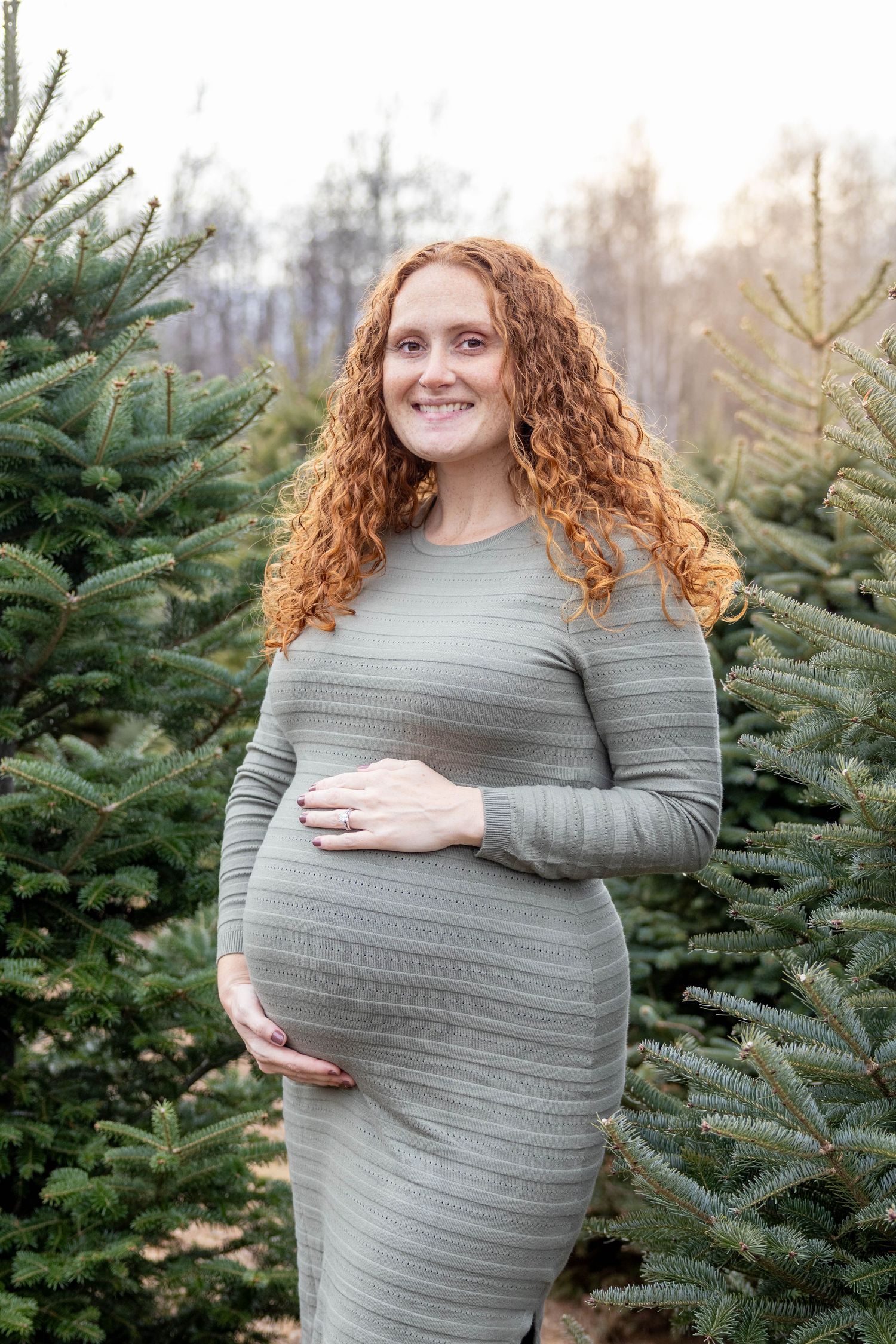 Pregnant woman in a sage green dress poses among evergreen trees at a Christmas tree farm.