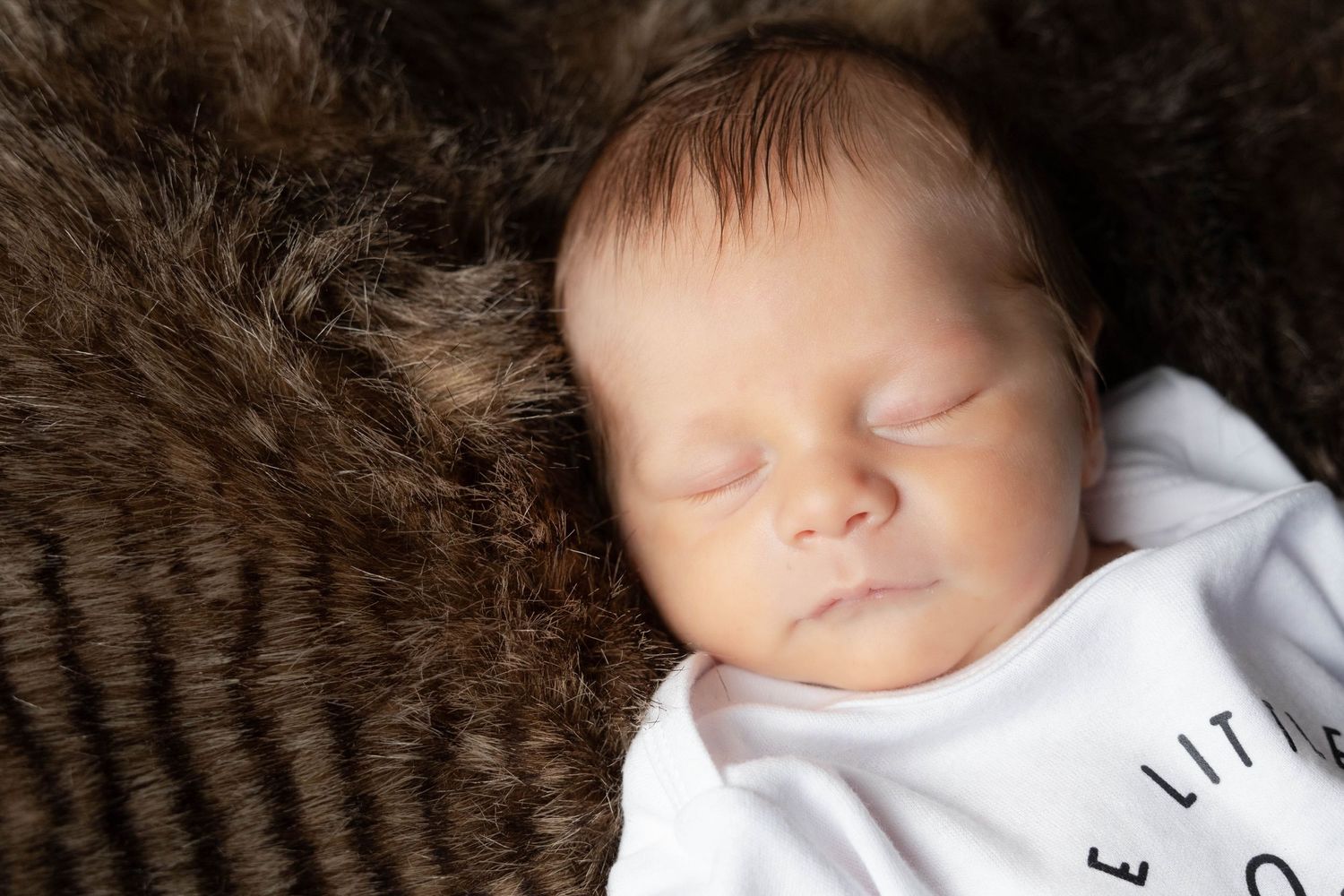 Sleeping newborn baby wearing white onesie on brown fur background.