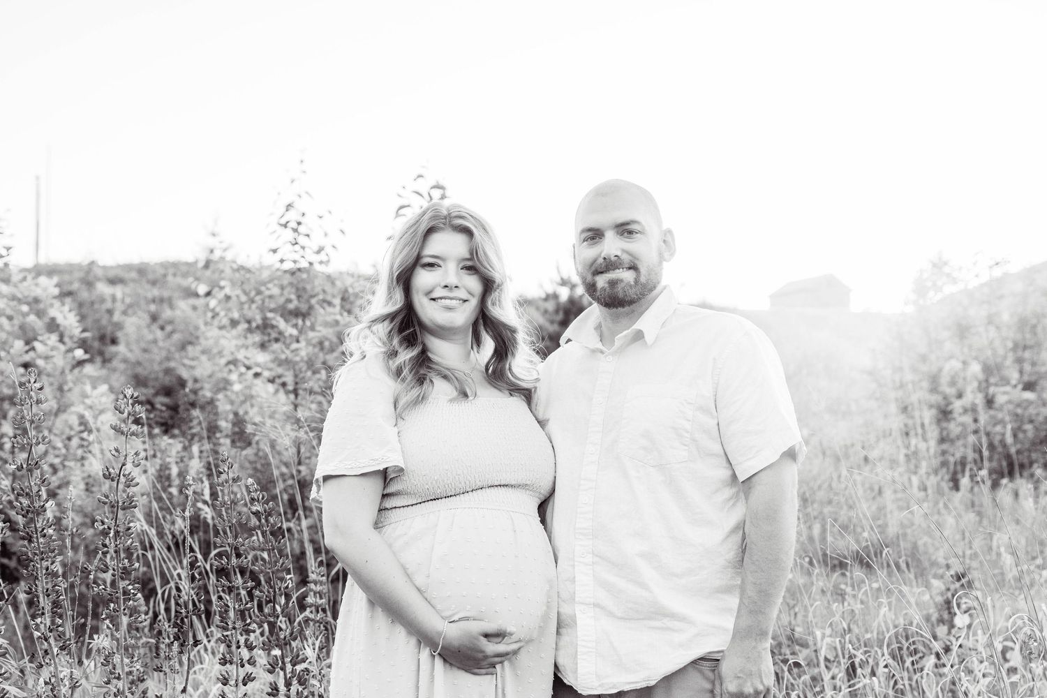 Black and white photo of an expecting couple posing together in a sunlit meadow during a maternity photoshoot.