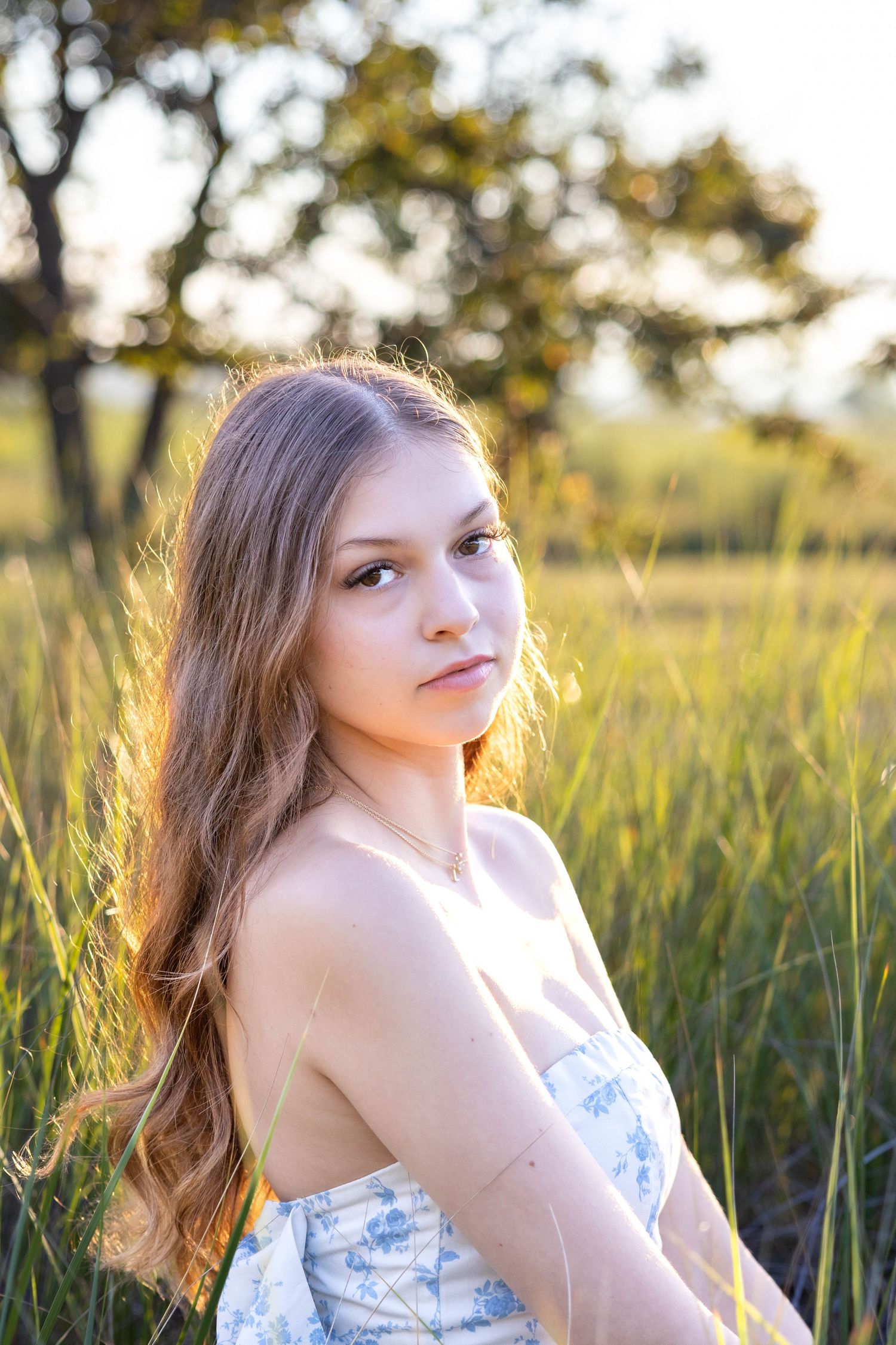 A woman in a floral dress poses thoughtfully in tall grass during golden hour lighting.