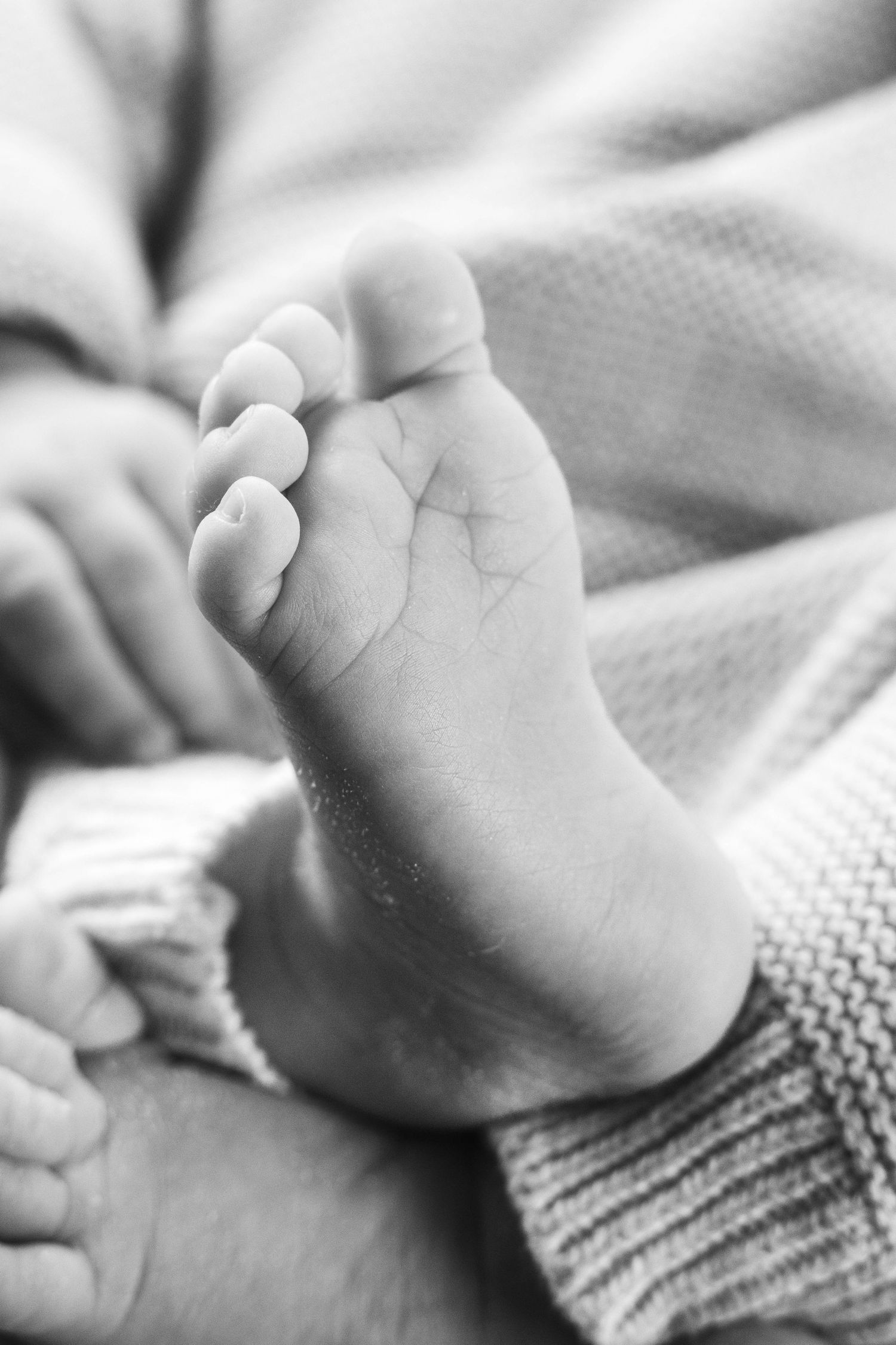 A close-up black and white photograph of a tiny baby foot resting on a textured knit blanket.