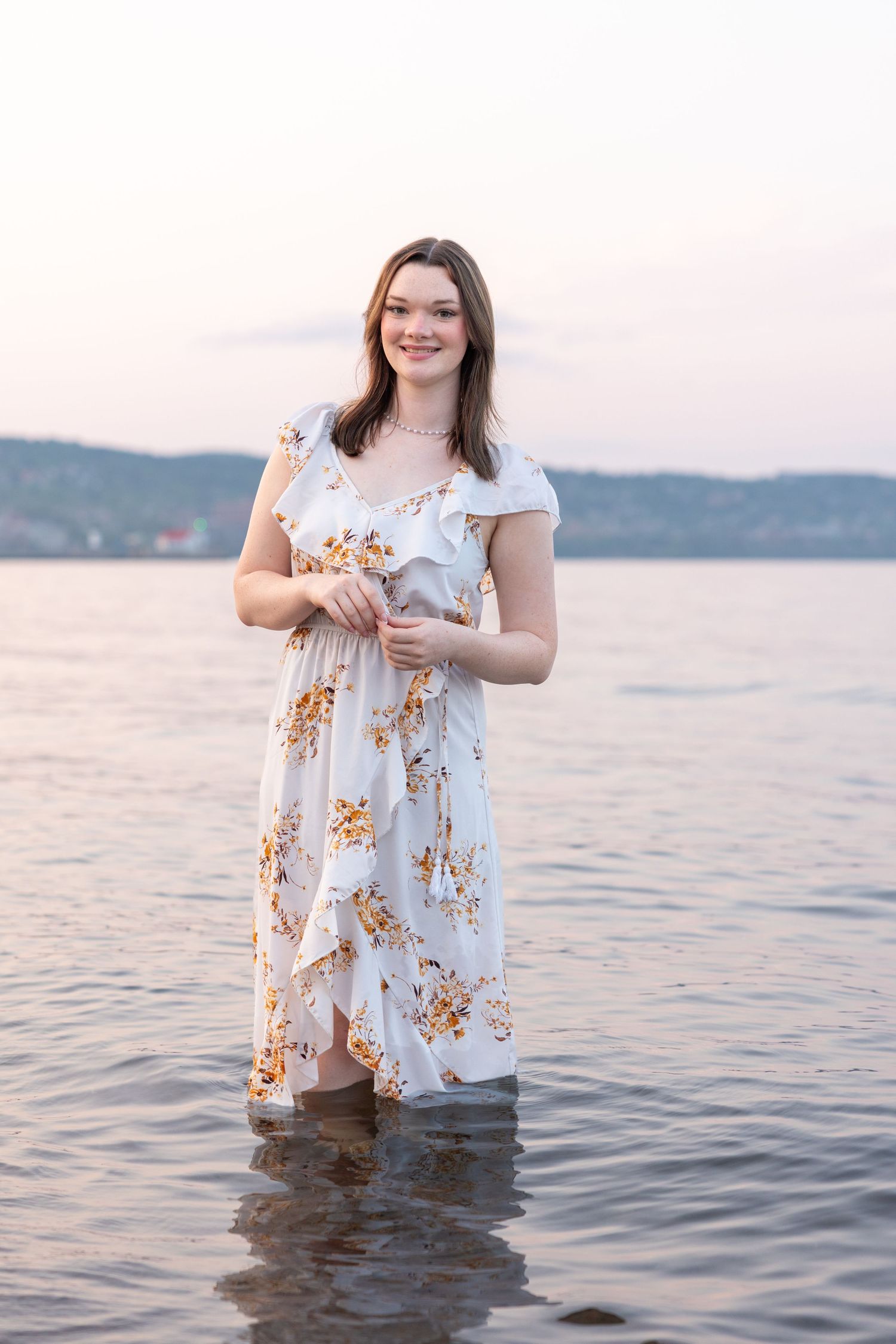Young woman in white floral maxi dress standing in shallow water at sunset, smiling gracefully.