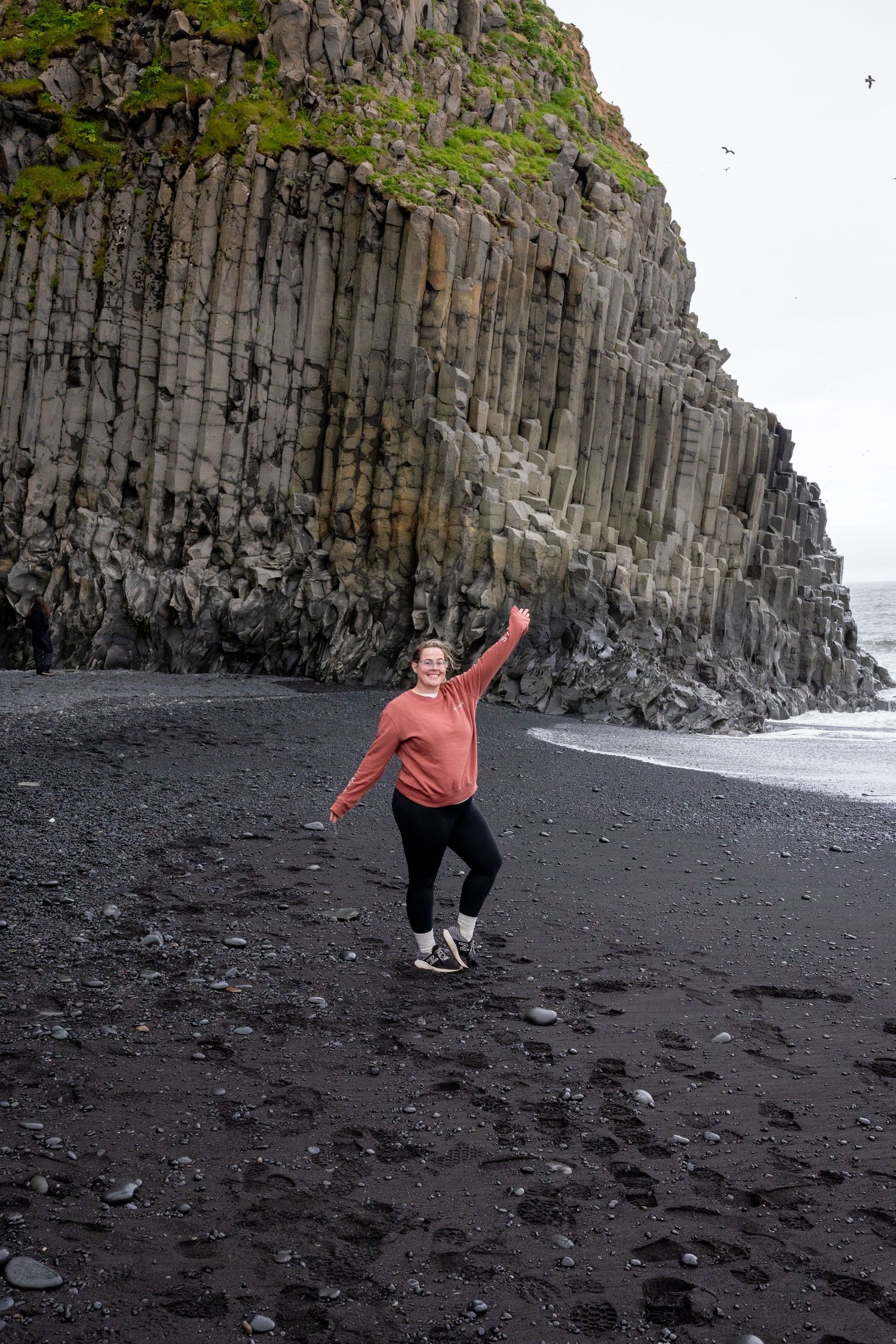 Woman in pink sweater poses on black sand beach at Reynisfjara, Iceland, with dramatic basalt column cliffs behind her.