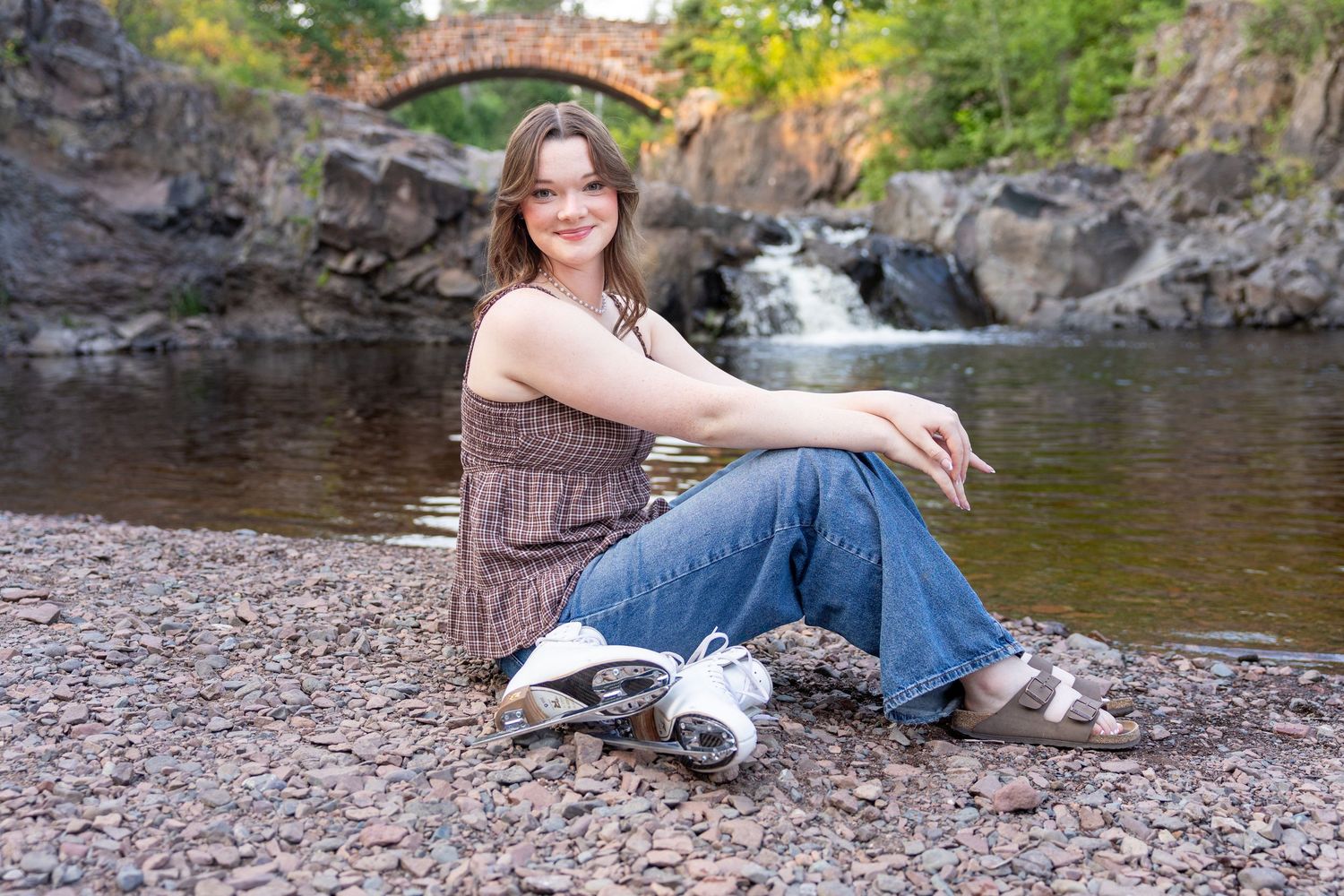 Young woman sitting on rocky riverbank smiling, with a stone arch bridge and waterfall in the background.