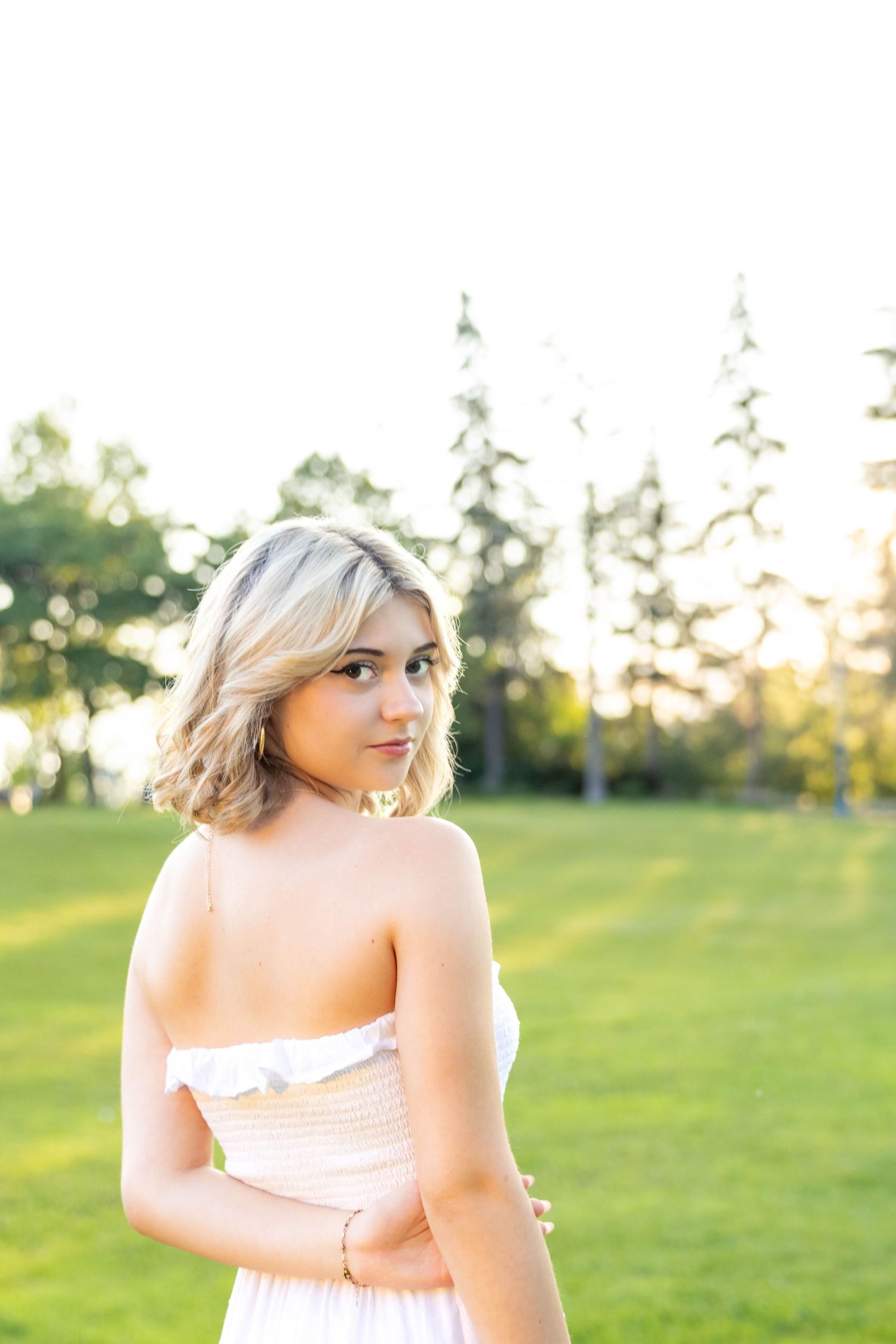 A woman in a white dress poses outdoors at sunset in a grassy field with evergreen trees in the background.