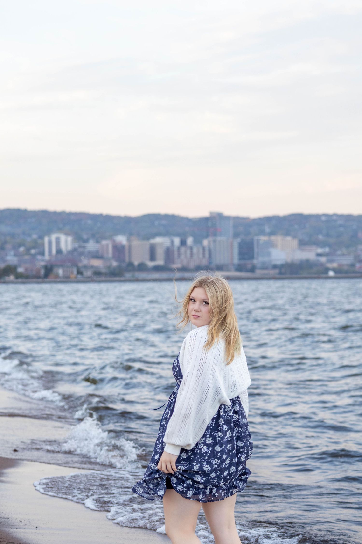 Woman in white sweater and floral dress walks along waterfront with city skyline in background at dusk.