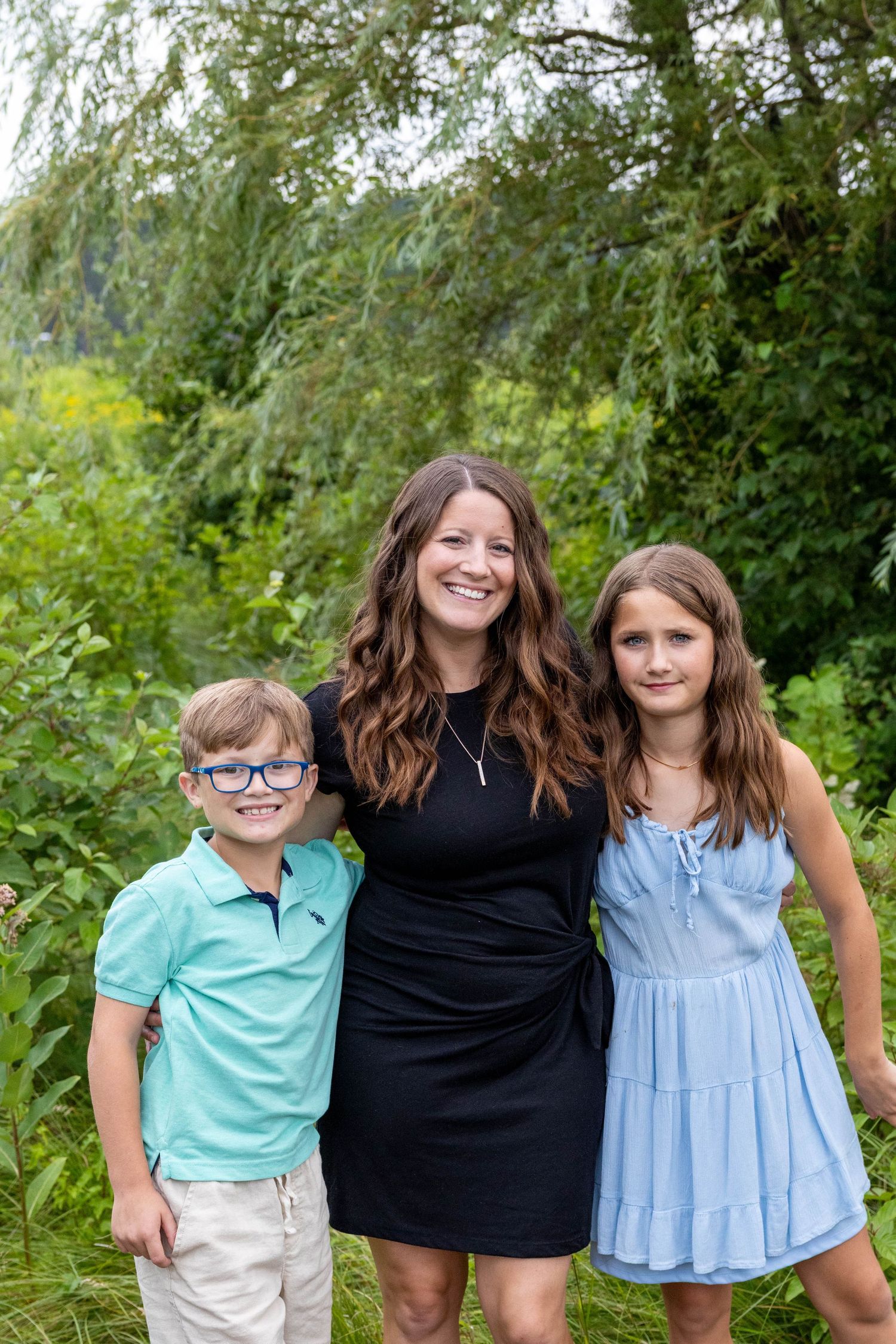 Three people stand together outdoors against a green leafy background in casual summer attire.