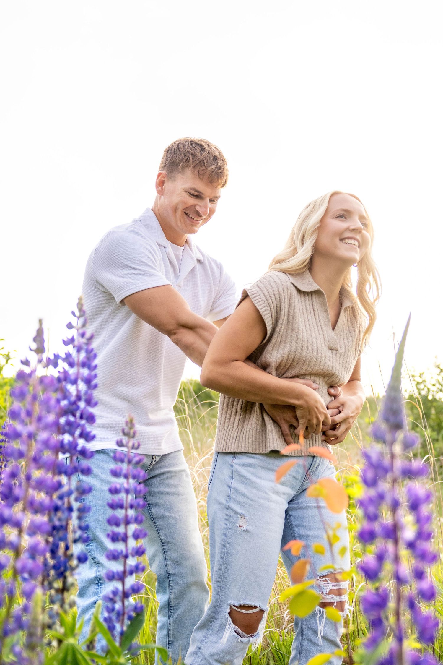 A joyful couple laughs together while standing in a field of blooming purple lupine flowers on a sunny day.