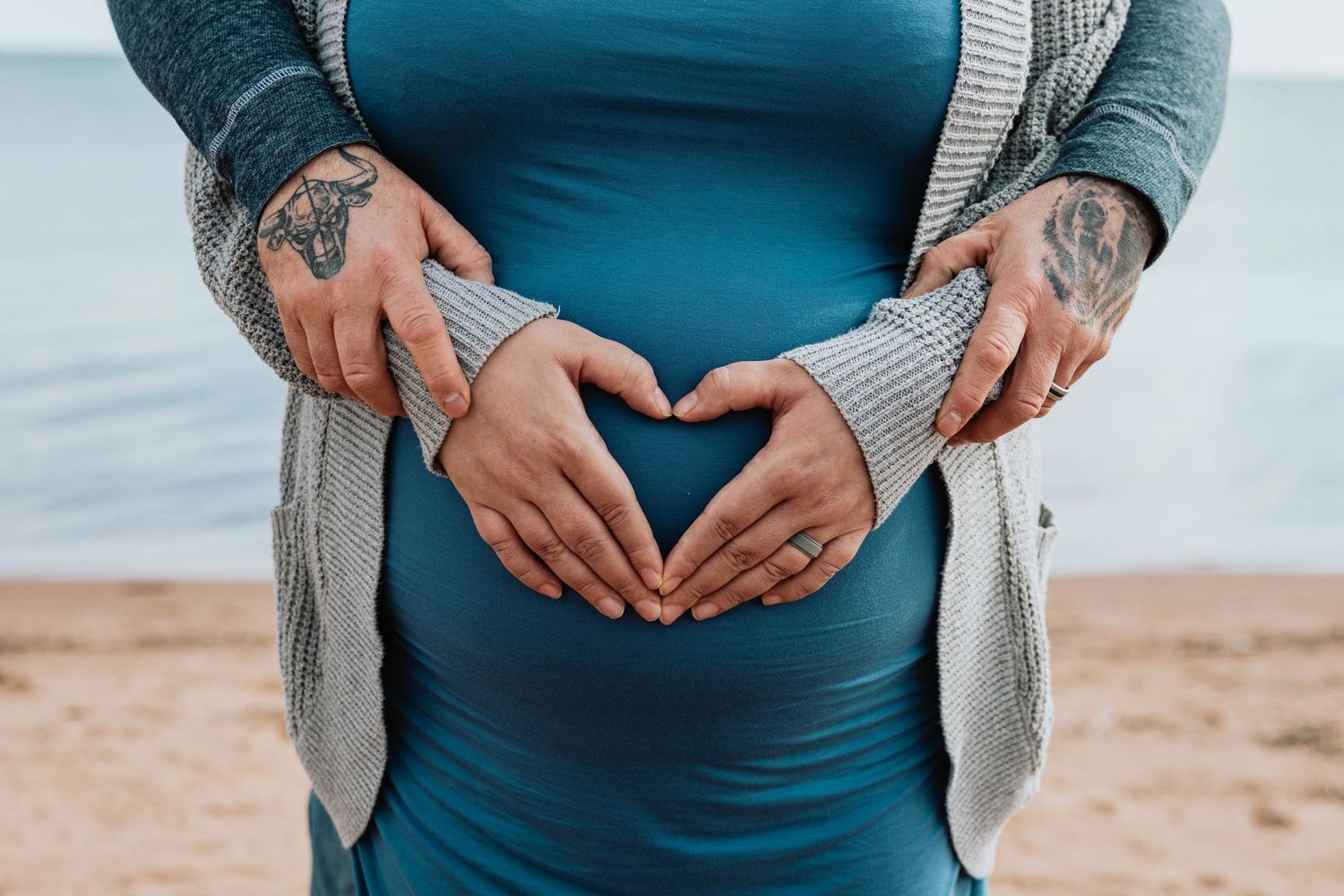 Tattooed hands form a heart shape over a pregnant belly in a blue dress. Park Point Duluth MN