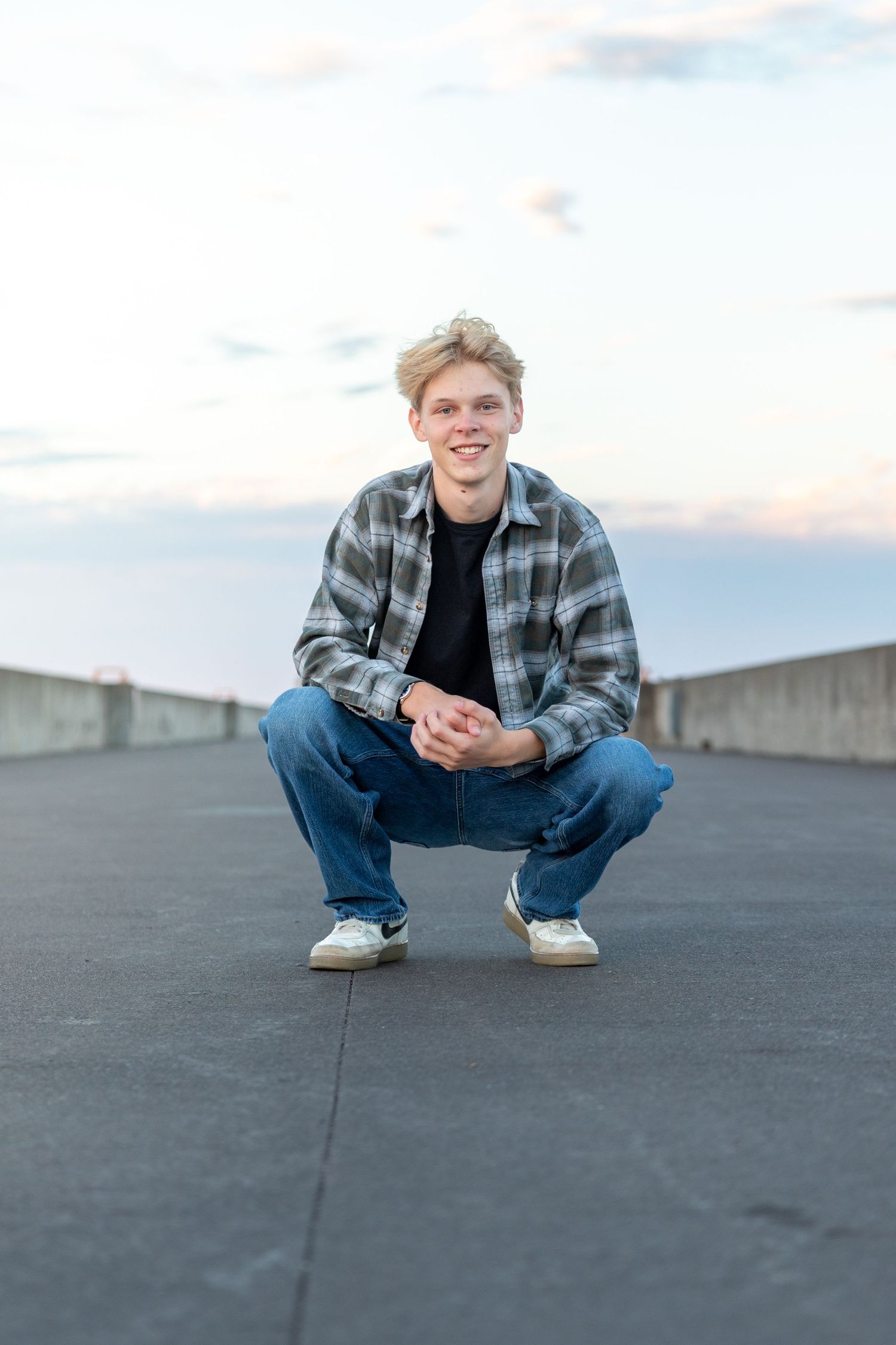 Young man in plaid shirt and jeans crouching outdoors, smiling confidently against a cloudy sky backdrop.