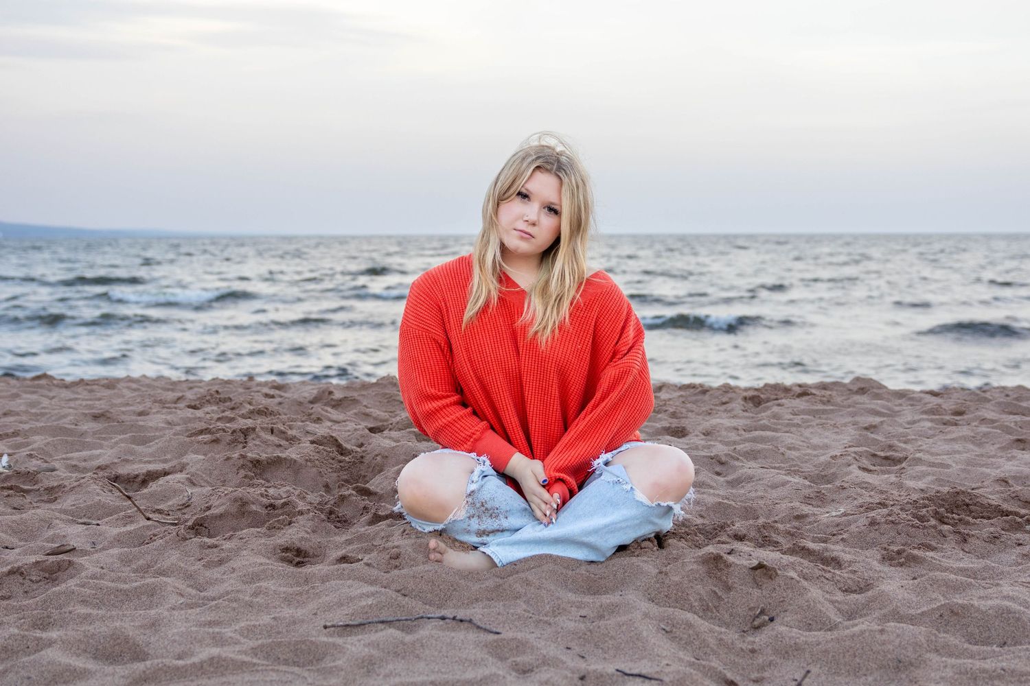 A person in a bright red sweater sits on a sandy beach near the ocean waves at sunset.