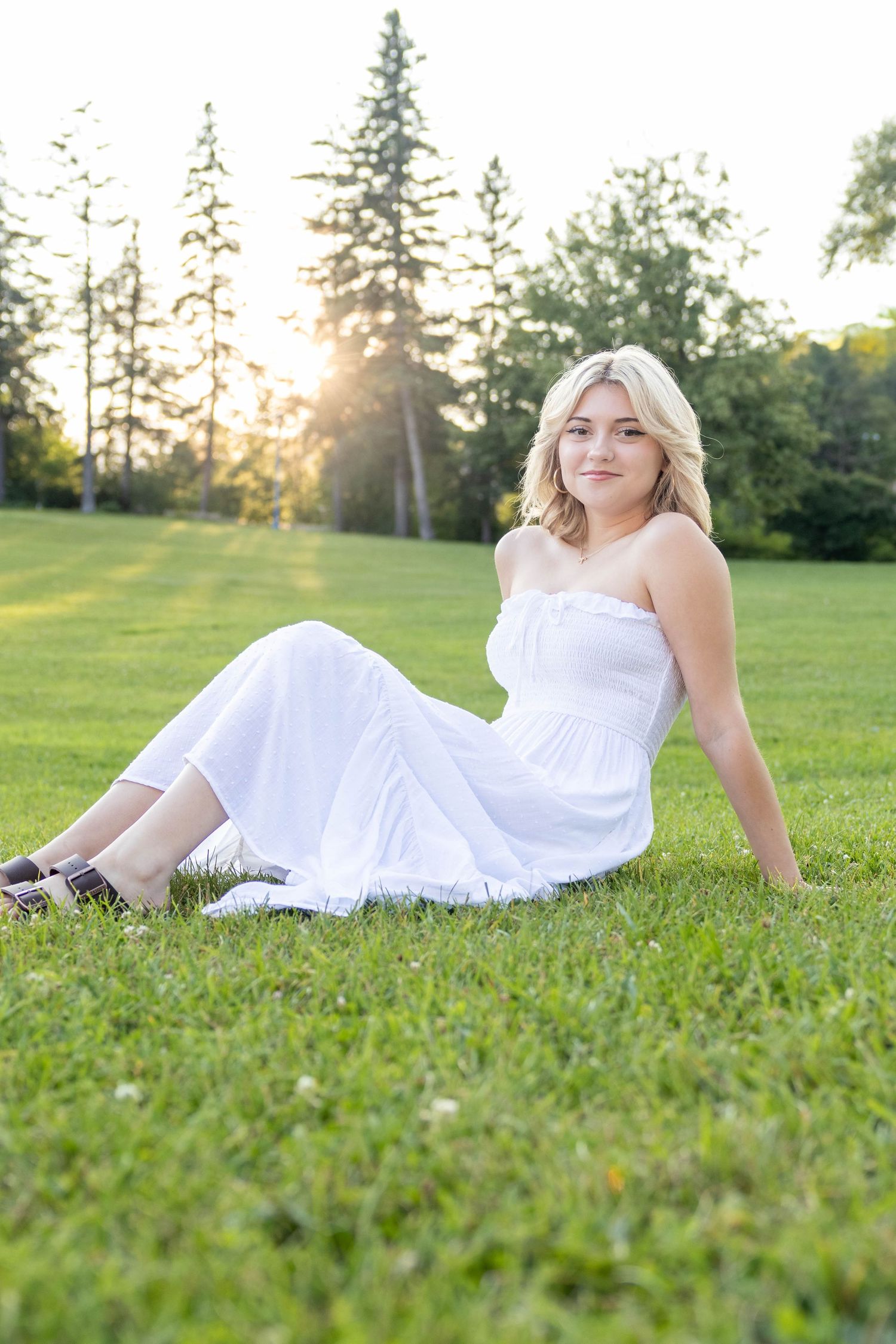 A woman in a white sundress reclines on green grass during golden hour with tall pine trees behind her.