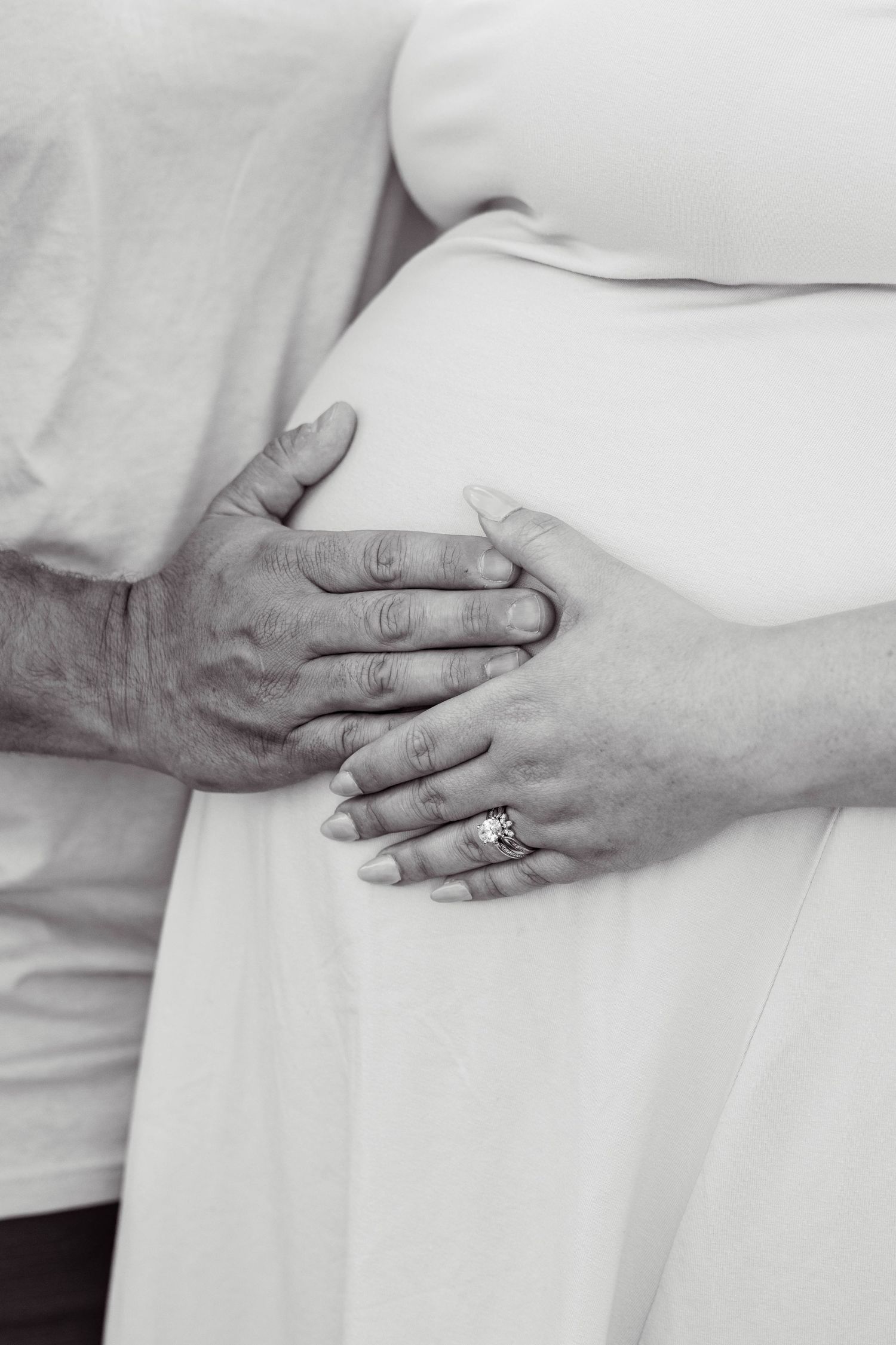 Black and white photo of hands clasped over a pregnant belly, with one hand wearing an engagement ring.