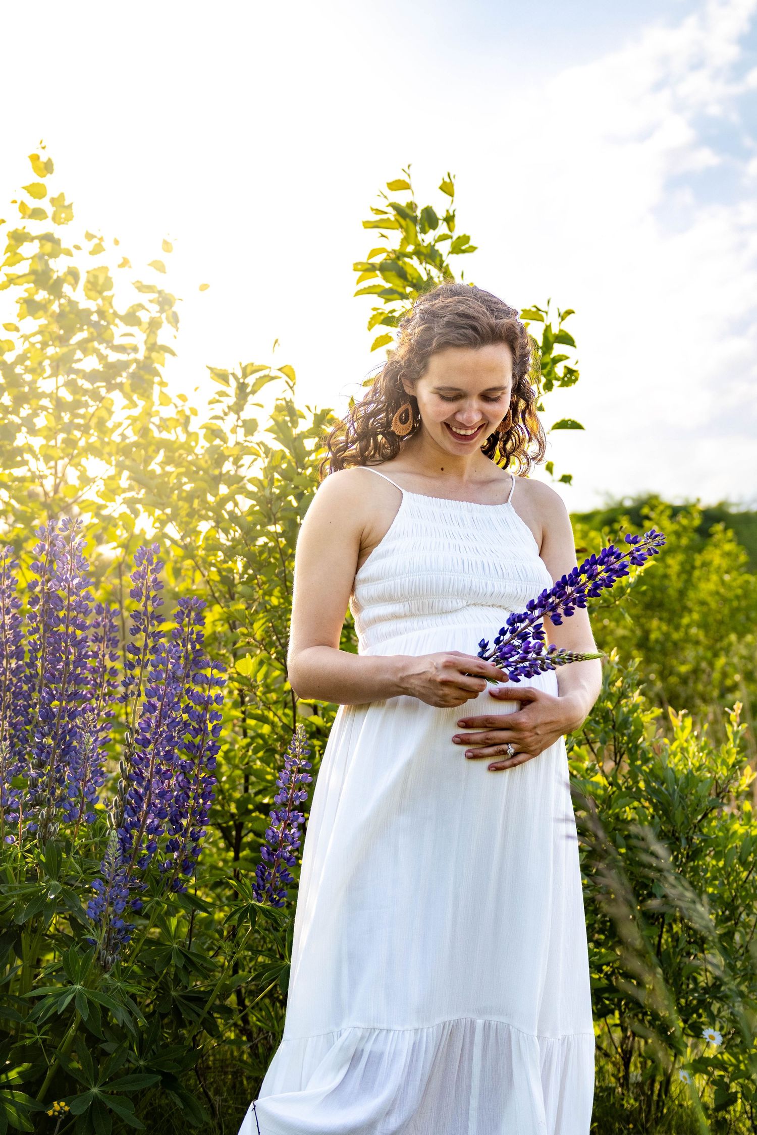 Woman in white sundress standing in garden holding purple lupine flowers on sunny summer day.