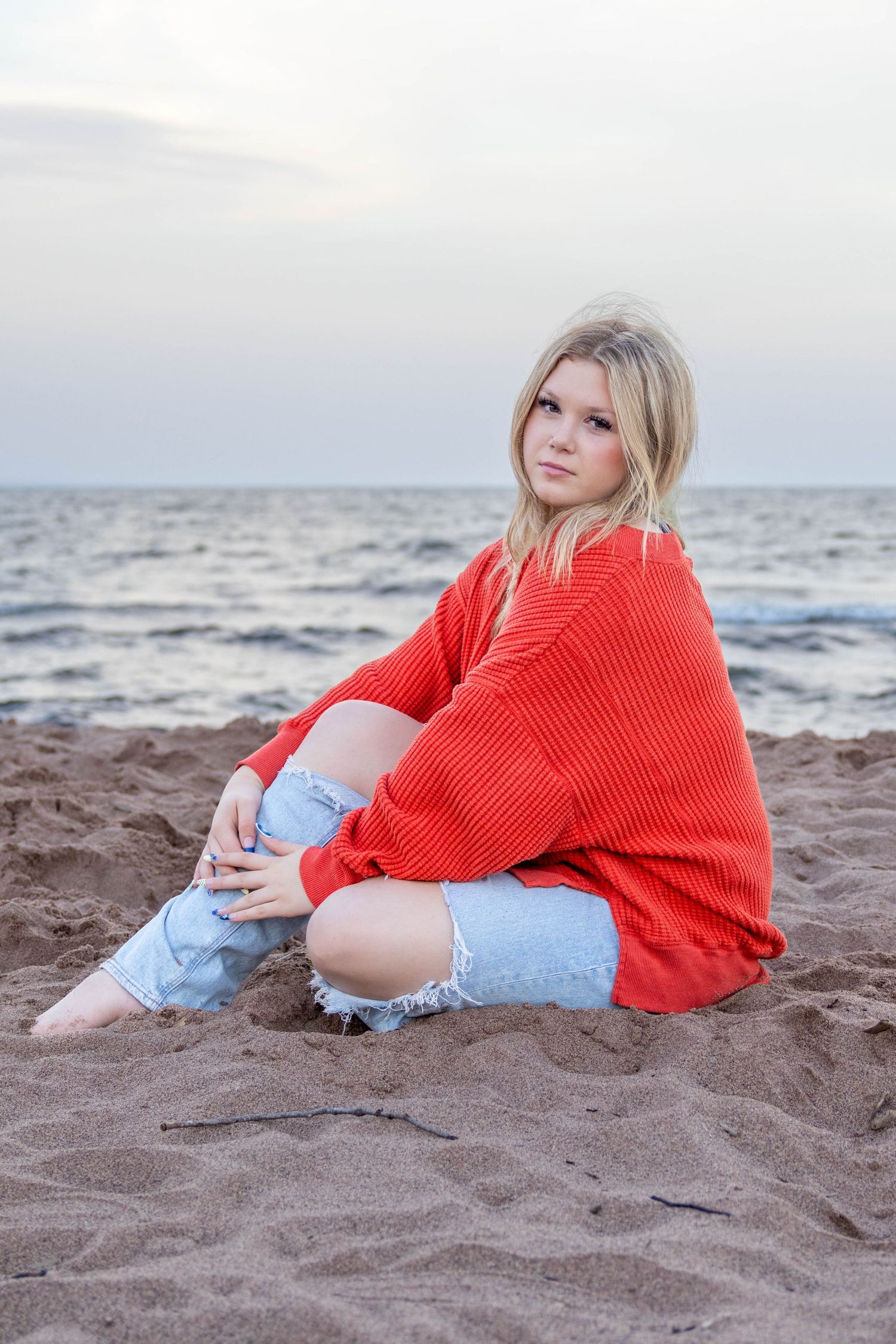 Blonde woman in red sweater and ripped jeans sitting on sandy beach by ocean at dusk.
