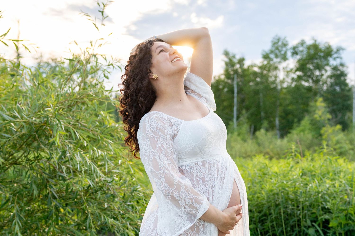Pregnant woman in white lace dress enjoying sunny day outdoors among green trees, cradling baby bump.
