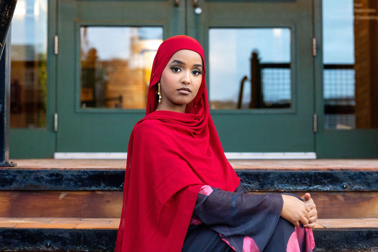A person wearing a bright red hijab and navy blue outfit sits thoughtfully on outdoor steps near a green door.