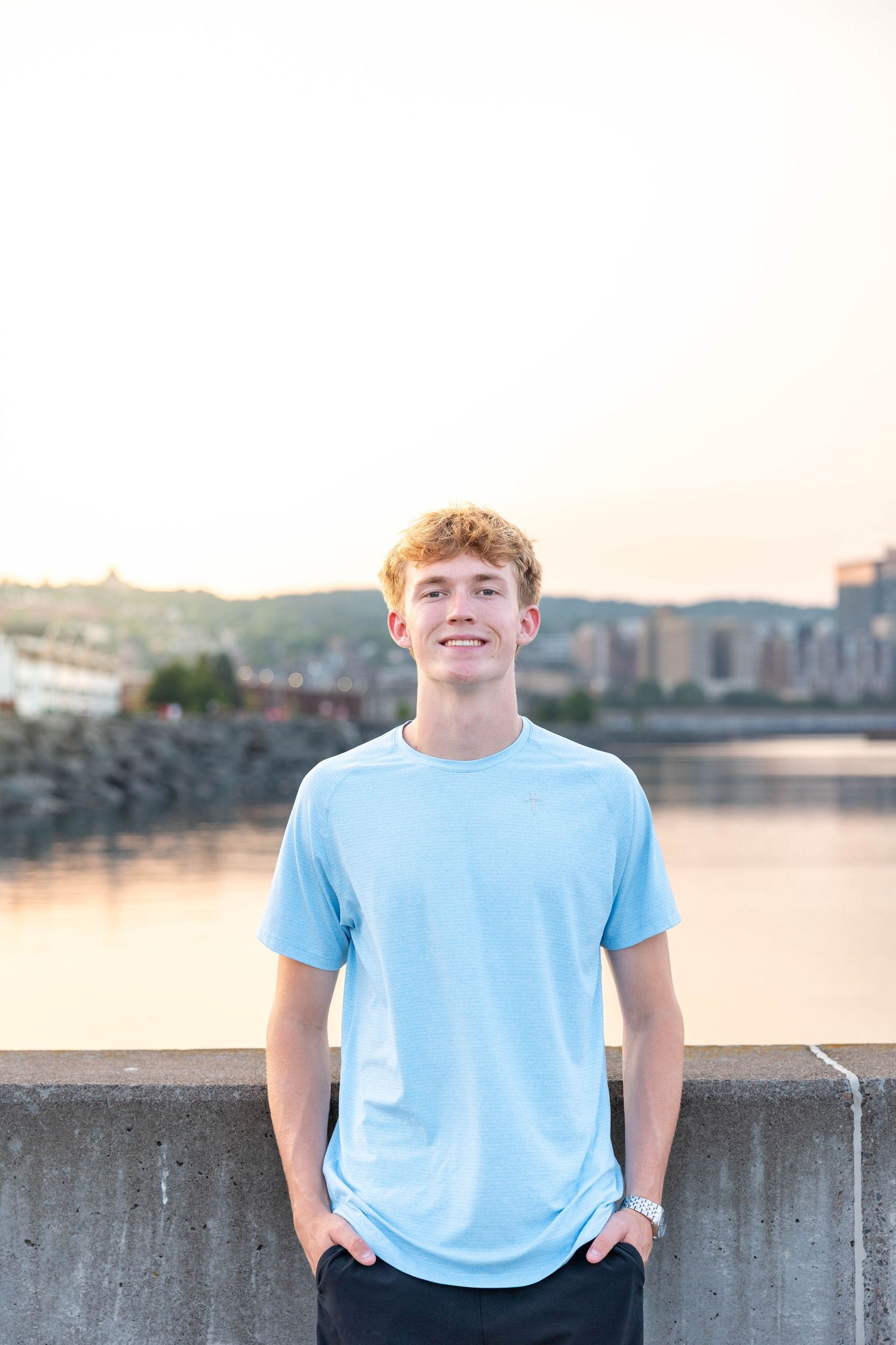 Young man in light blue t-shirt smiling outdoors by a river at sunset with city skyline in background.