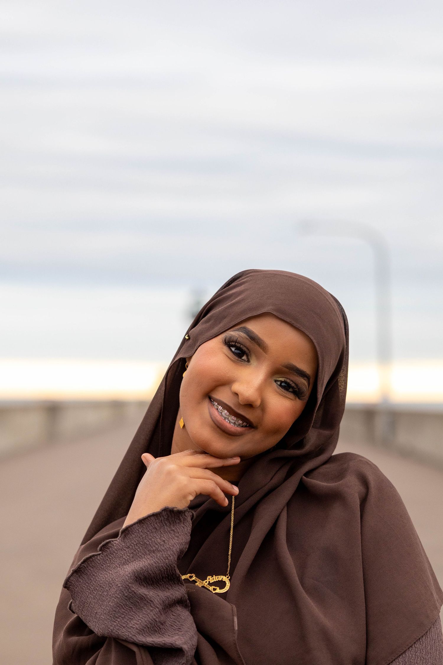 Person in brown hijab and abaya poses outdoors with a bright smile.