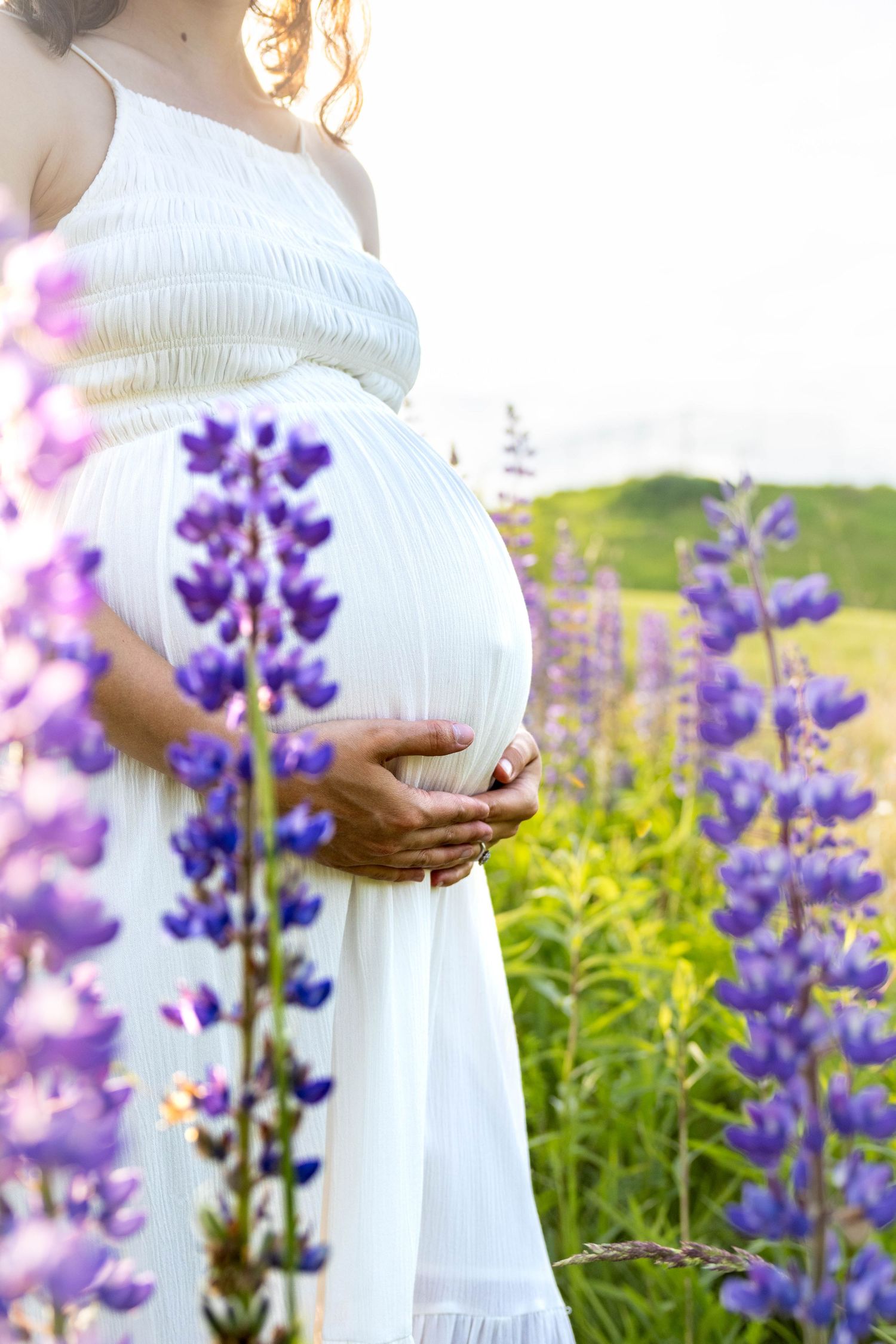 Person in flowing white dress stands in a garden of vibrant purple lupines and yellow wildflowers on a sunny day.
